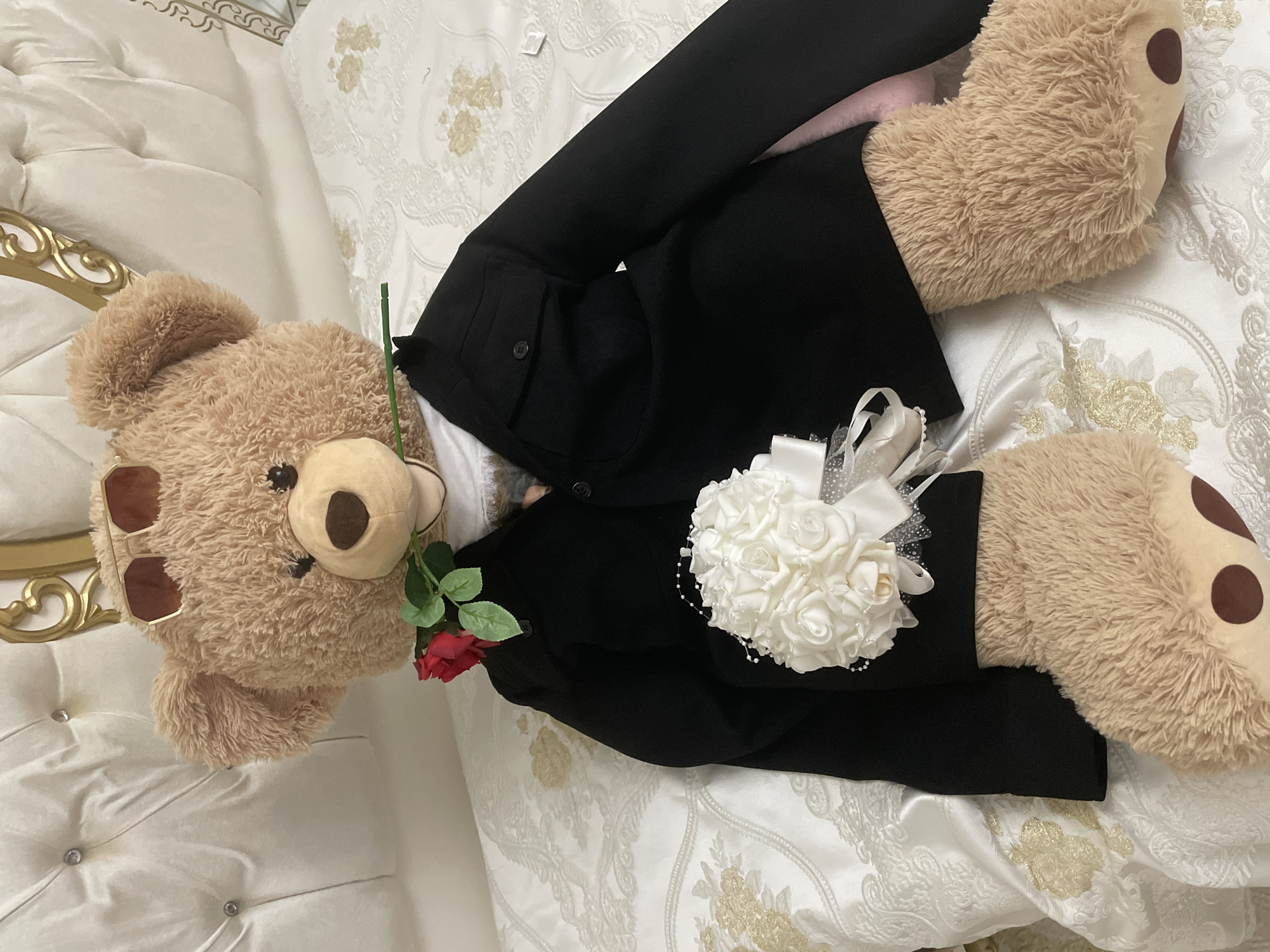 The image displays a large, light brown teddy bear formally arranged on a luxurious bed. The teddy bear is dressed in a black suit jacket and wears brown-tinted sunglasses pushed up onto its head. It holds a single red rose with green leaves in its left paw. Attached to its right arm or lapel is an elegant white boutonnière or corsage, fashioned from multiple small white artificial roses, adorned with white satin and sheer ribbons, and delicate pearl strings.
The teddy bear rests on a white bedspread featuring an intricate, possibly embroidered or jacquard, pattern in gold and white. A portion of a plush, tufted white headboard or bed frame, embellished with decorative silver or crystal buttons and an ornate gold-painted trim, is visible on the left. The overall scene suggests an indoor setting, likely a well-appointed bedroom, arranged for a romantic gesture or a special occasion such as a wedding. There are no indications of time of day, weather, or any visible text. This media file is associated with the location of Ihtiman, Bulgaria.