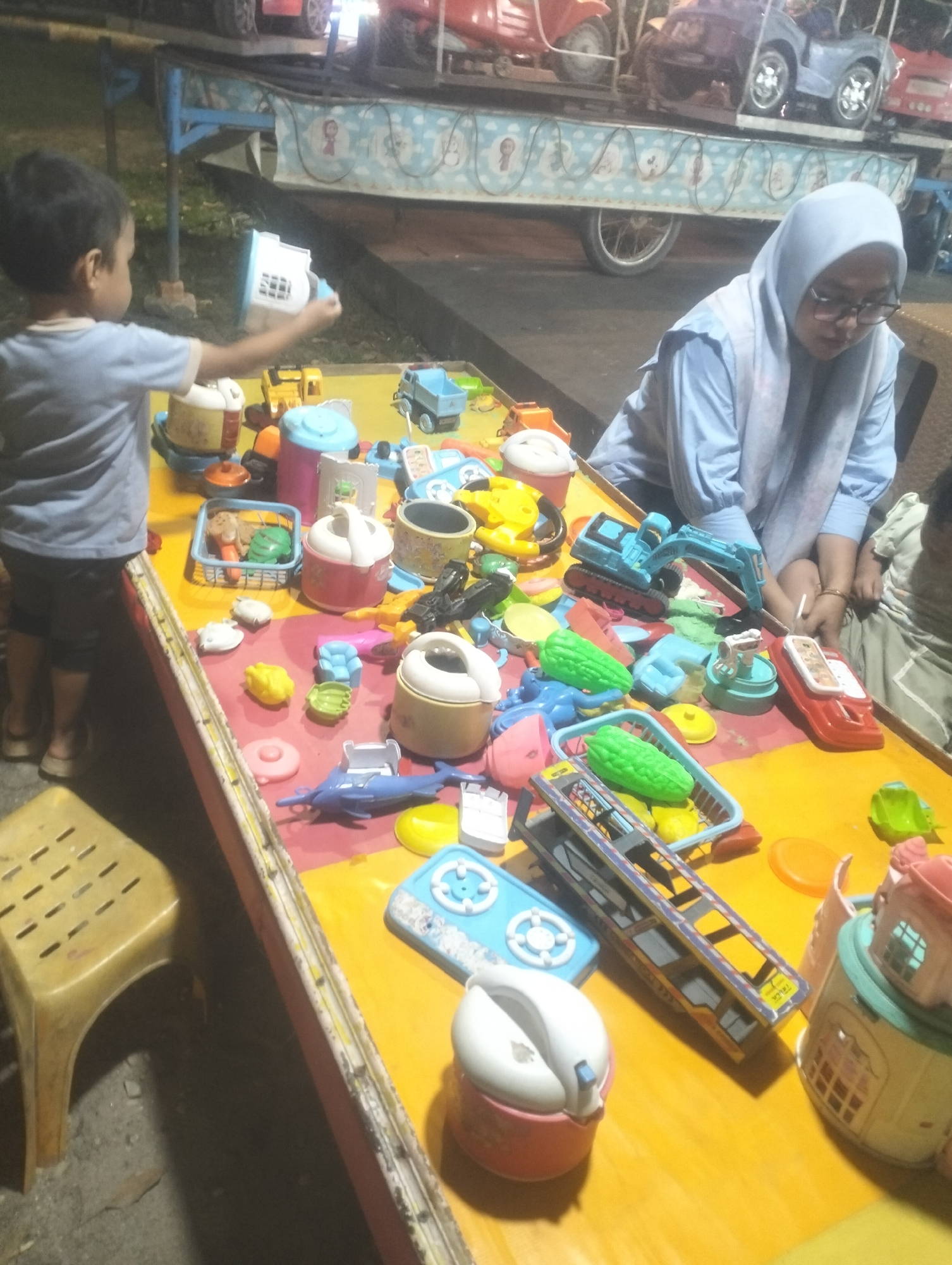 A young boy in a blue t-shirt and dark shorts is holding a white and blue toy in his outstretched right hand. He is standing to the left of a long, yellow table filled with various toys. To the right of the boy, a woman wearing a light blue hijab and glasses is seated and looking down at the table. The table is covered with a collection of plastic toys including construction vehicles, miniature houses, food-shaped toys, and buckets. In the background, there are more toys on display, possibly at a playground or an outdoor toy market in Dumai City, Indonesia. The ground appears to be rough concrete.