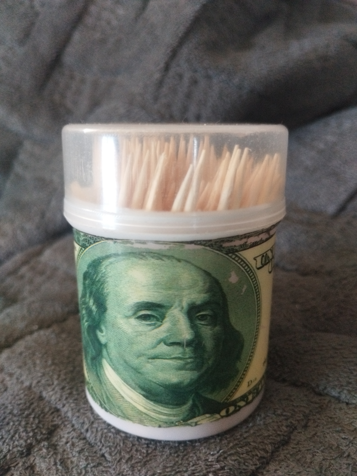 The image shows a close-up, eye-level, indoor shot of a plastic container filled with wooden toothpicks. The container is cylindrical and decorated with a repeating pattern resembling a U.S. one-hundred-dollar bill, featuring a portrait of Benjamin Franklin. The background is a dark gray, textured fabric, likely a blanket or upholstery. The lighting is soft and diffused, coming from above and slightly to the right. There are no people or animals visible in the image, and no specific location in Oskemen, Kazakhstan is discernible from the provided visual information.