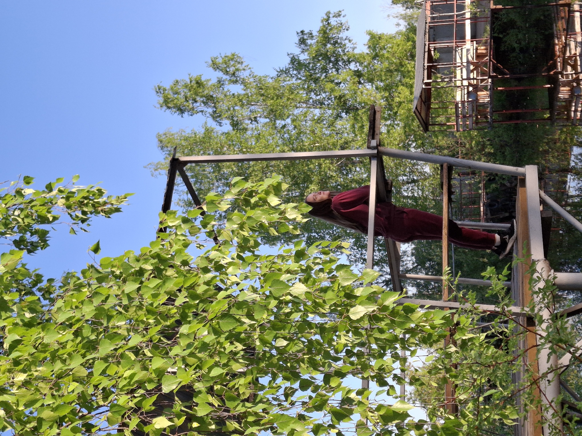 The image depicts a person lying atop a metal structure, appearing to be part of a playground or recreational area. The person is wearing a dark red outfit, possibly a jumpsuit or similar attire, and dark shoes. They are positioned horizontally on the structure, with their head near the center and their body extending towards the right.
The metal structure consists of pipes and supporting beams, with signs of rust and weathering. A dense canopy of green leaves and branches surrounds the structure, partially obscuring the view. In the upper right corner, the skeletal frame of another structure, possibly a building or a climbing frame, is visible.
The background reveals a clear blue sky, with some treetops visible. The image seems to be taken from a low angle, looking upwards towards the subject and the surrounding structures. Based on the information provided, the location is Chaikovsky, Russia.