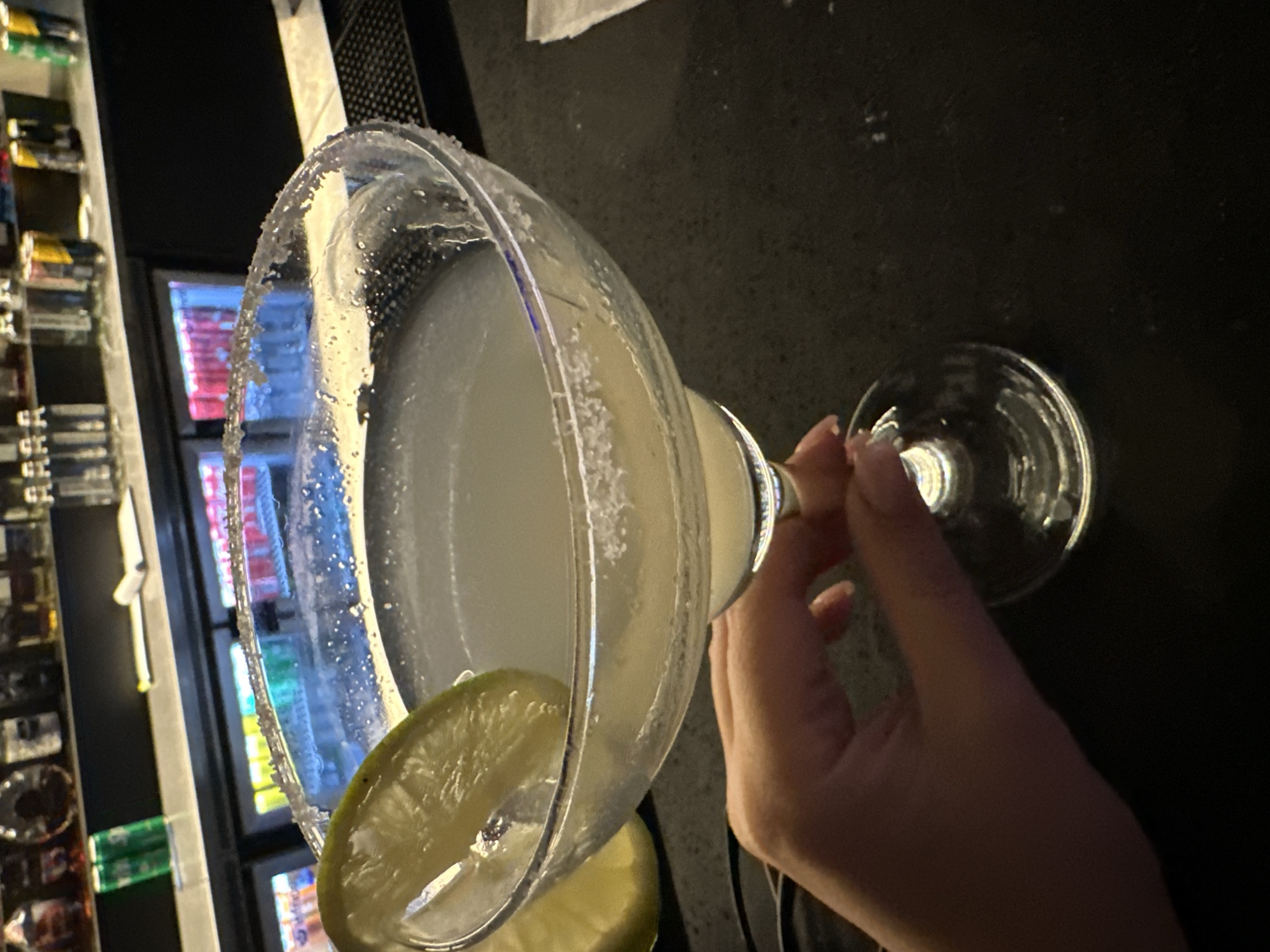 A person's hand holds a margarita glass with a salted rim, garnished with a lime wedge, in what appears to be a dimly lit bar or restaurant setting. The glass contains a light-colored beverage, typical of a margarita. The hand, with neatly manicured nails, grips the stem of the glass. In the background, shelves stocked with various bottles or cans are visible, along with glowing, colorful display screens, possibly from a cooler or TV, indicating an indoor commercial establishment. The dark surface beneath the glass suggests a bar counter. The overall impression is of an evening or night out, with the subject enjoying a drink. No discernible text is visible in the image.