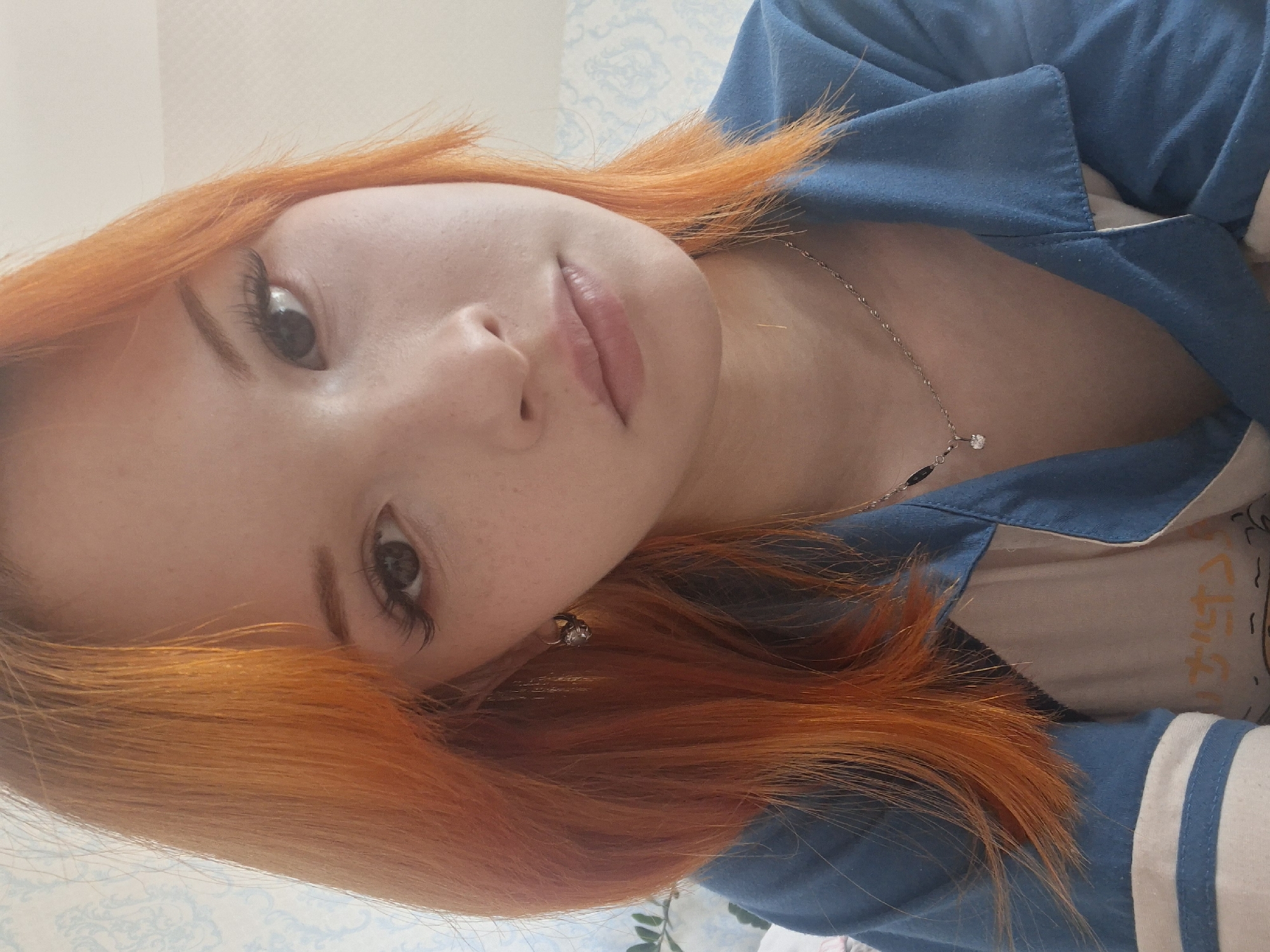 A close-up portrait of a fair-skinned woman with striking orange hair. Her gaze is directed towards the camera. She is wearing a blue collared shirt over a light-colored top. A delicate silver necklace adorns her neck. Freckles are visible on her face. The background suggests an interior setting with a patterned wallpaper. The location is identified as the city of Chaikovsky, Russia.