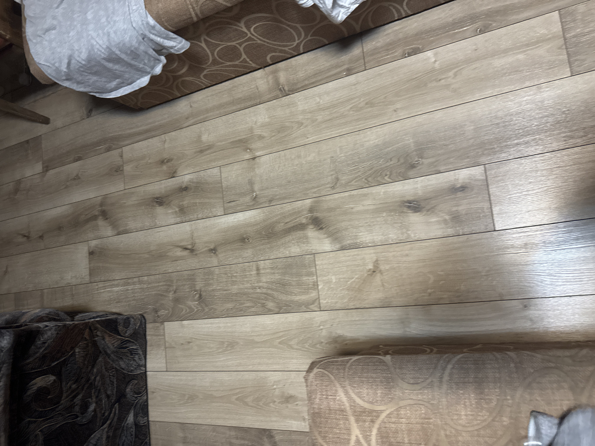 The scene is an interior shot of a room, possibly in a home. The floor is covered with light-colored wood-look planks. There are parts of furniture visible in the image. A brown armchair with a pattern is in the upper portion. In the lower left is a dark brown patterned upholstered item, likely furniture. In the lower right corner is another tan-colored patterned piece of furniture. No people are visible. The location is unknown, not specified as Lehchevo, Bulgaria.