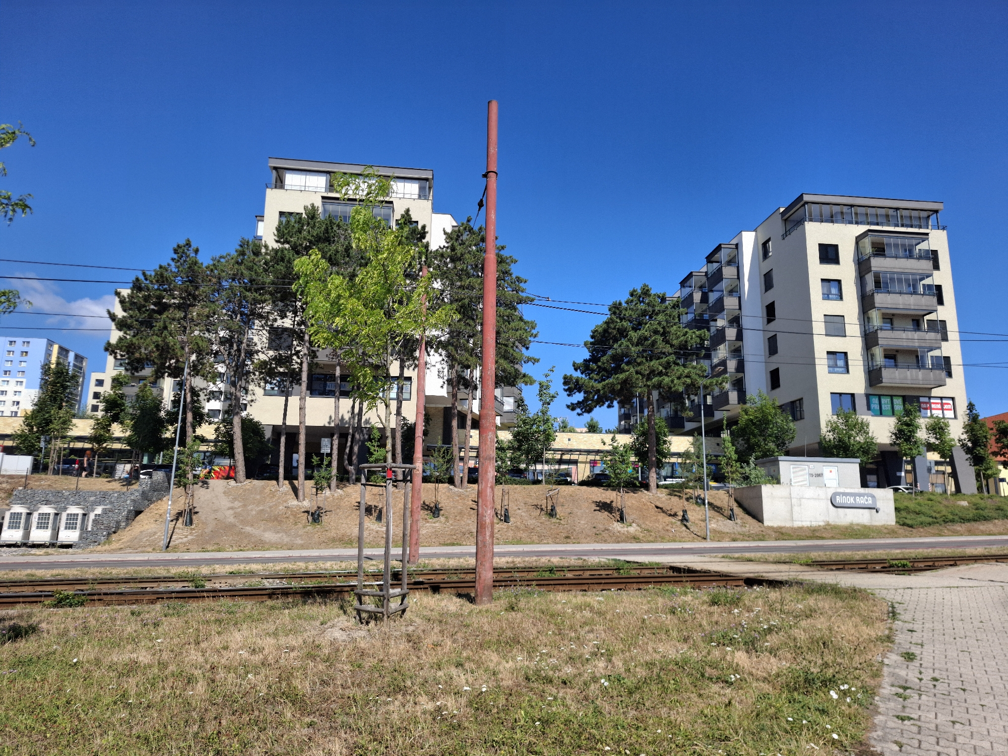 The image shows a clear, sunny day in Bratislava, Slovakia. In the foreground, there are train tracks and dry grass. Behind the tracks are two modern apartment buildings with balconies. To the left of the apartment buildings, there is a blue building. A red pole stands in the middle of the image, in front of the apartment buildings. A sign with the text "RÍNOK RAČA" is visible on the right side of the image. The overall scene is urban, with a mix of residential buildings and public infrastructure.