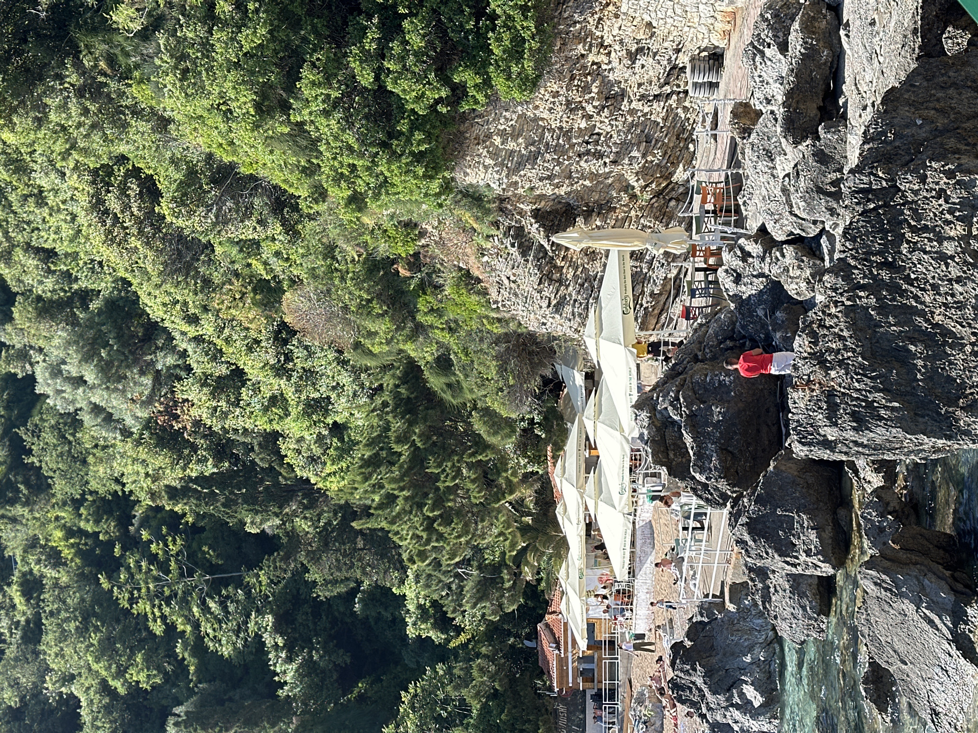 The image shows a coastal scene in Budva, Montenegro. Lush green vegetation covers a steep hillside on the left. A rocky cliff forms the right side of the frame. In between the cliff and hillside, a beach area is visible, populated with people and shaded by numerous white parasols. People are seen walking and relaxing on the beach. One person in a red shirt stands on the rocks. The turquoise water of the Adriatic Sea is seen at the bottom of the frame. A structure on the hillside appears to be a restaurant or bar, indicated by tables and chairs beneath the parasols.