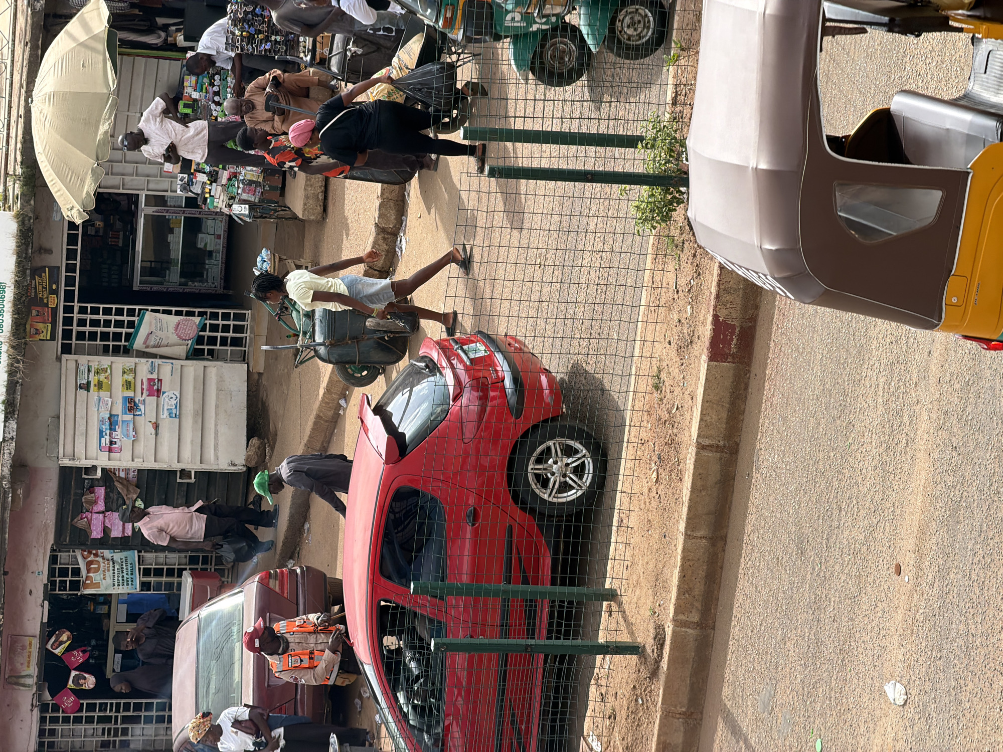 The image depicts a busy street scene in Bukuru, Nigeria, likely a market area. In the foreground, a red Peugeot hatchback is parked. A wire mesh fence runs along the street. Behind the fence, a group of people are engaged in various activities. Some are standing or walking, while others are seated. There are several stalls with goods displayed, including what appears to be clothing and accessories. A woman in a white shirt and headscarf is standing near a maroon sedan. A person in an orange vest and red cap is also visible. A green wheelbarrow is present, as is a three-wheeled auto rickshaw (Keke Napep) in the far right foreground. Buildings line the street, some appearing to be shops with awnings and open entrances. The ground is paved with asphalt and dirt. The overall impression is one of a vibrant, commercial district.