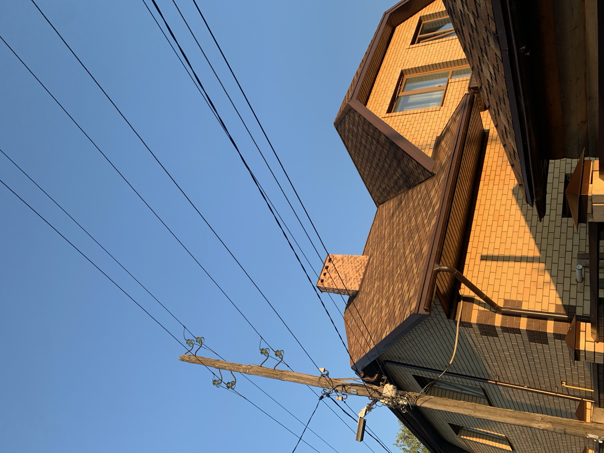 The image shows a two-story house with a brick exterior, brown asphalt shingle roof, and dark brown gutters. A brick chimney is visible on the roof. The house is illuminated by the warm, golden light of the setting sun, casting long shadows. Several overhead power lines crisscross the clear blue sky, supported by a wooden utility pole with insulators. The pole also has some electrical equipment attached, including a street lamp fixture. A portion of a fence made of brick and dark wood is in the foreground. The scene is likely a residential area. This image was taken in Asha, Russia.
