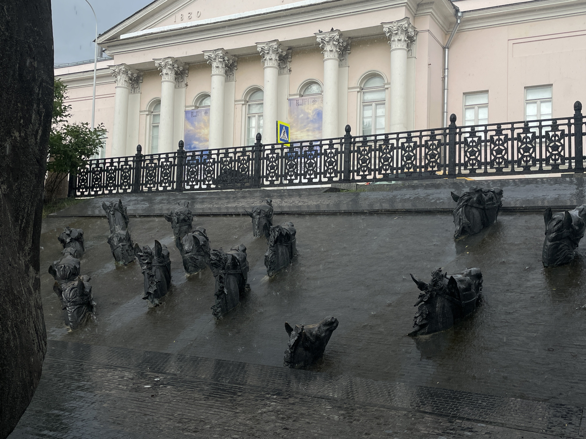 The image captures a wet, overcast day in Moscow, Russia, likely during a rain shower, as evidenced by the falling raindrops and wet surfaces. In the foreground, a series of dark, textured sculptures are partially submerged in water on a paved surface. These sculptures appear to depict horse heads and parts of horse-drawn carriages, arranged in a scattered formation. The rain has created ripples and puddles around them, reflecting the gloomy sky.
Behind the sculptures, a black wrought-iron fence with intricate patterns runs horizontally across the mid-ground, separating the sculpture area from the background. A yellow pedestrian crossing sign is visible above the fence. The background is dominated by a large, pale pink building with a neoclassical facade. It features a row of tall, white Corinthian columns supporting a pediment, and several arched windows. The year "1830" is inscribed above the central columns. The building's architecture suggests it is a significant public structure, possibly a museum or government building. The overall impression is one of somber weather conditions over a historical urban setting.