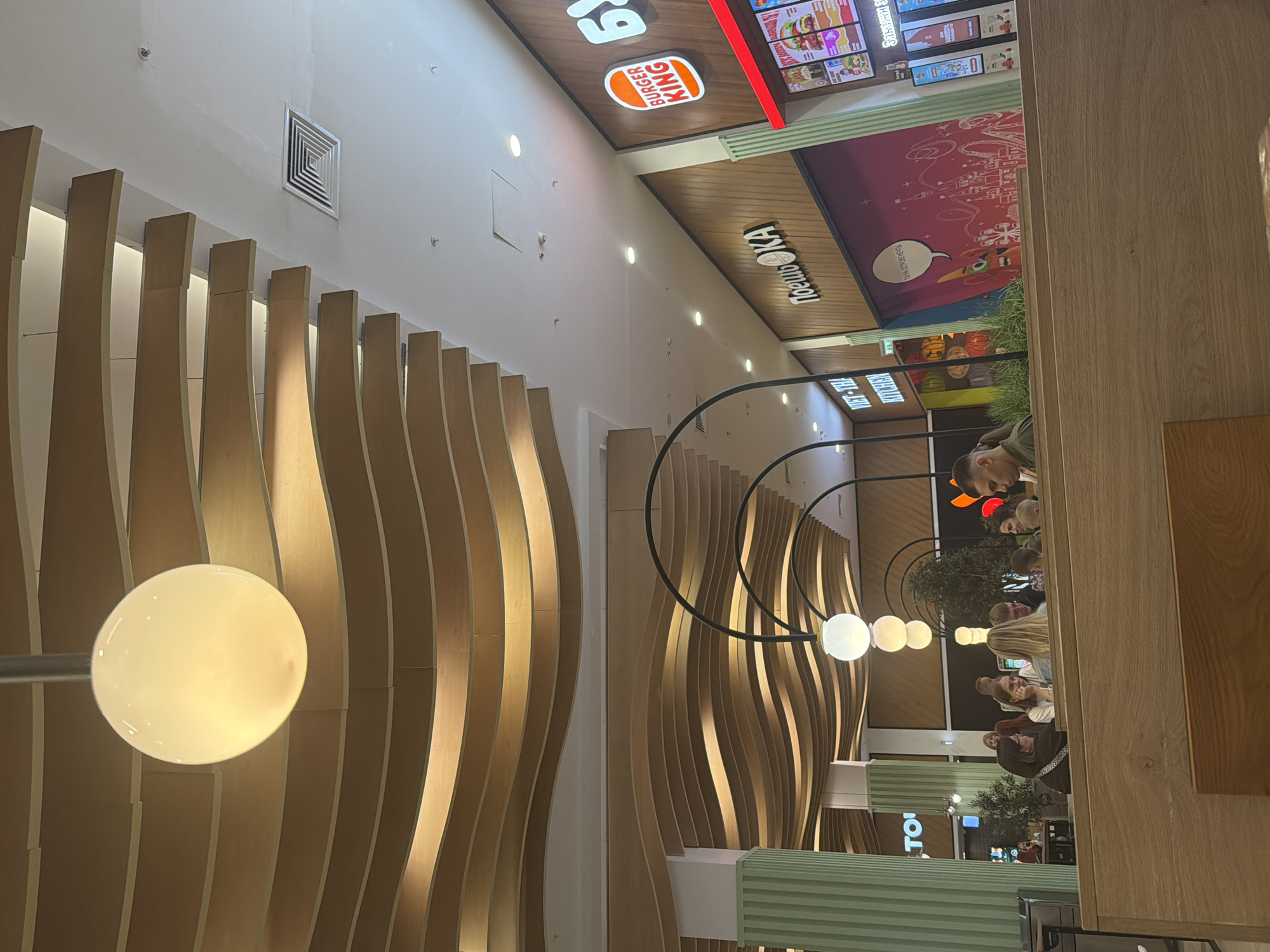 The image depicts an interior scene, likely a food court or a restaurant within a mall, with several people seated at tables. The overall atmosphere appears casual and relaxed. The setting features a modern design with curved wooden panels and prominent lighting. In the foreground, a large, circular light fixture with a diffused glow is visible.
To the left, the curved wooden panels create a wave-like pattern, illuminated from behind, giving a warm and inviting ambiance. In the background, a Burger King restaurant is clearly identifiable with its signage. Various menu boards are displayed above the counter. Further into the background, another sign reads "ТОСМО KA" in large, bold letters.
Several groups of people are seen dining or conversing at tables. Some individuals are looking towards the camera, while others are engaged in conversation. The lighting suggests it could be evening or a dimly lit indoor space. The presence of the Burger King and what appears to be other food establishments implies a public space designed for dining and recreation. The overall impression is that of a busy but comfortable dining area.