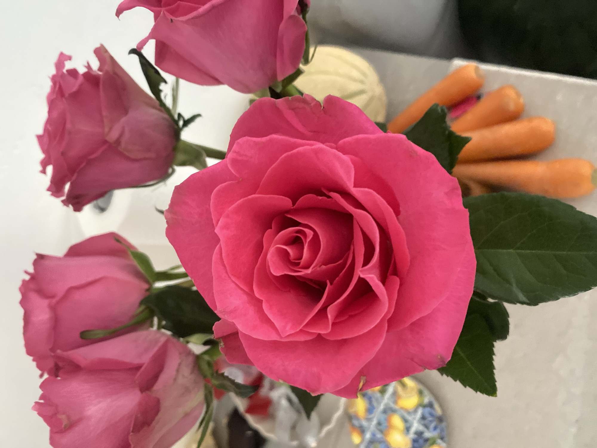 The image shows a bouquet of vibrant pink roses with green leaves. In the background, partially visible, are some small carrots. The roses are arranged in a vase that sits on a light-colored surface, possibly a countertop. The background is out of focus, showing hints of a wall and possibly other decorative items. The scene appears to be an indoor setting. There is no indication of the city of Lütjenwestedt, Germany.
