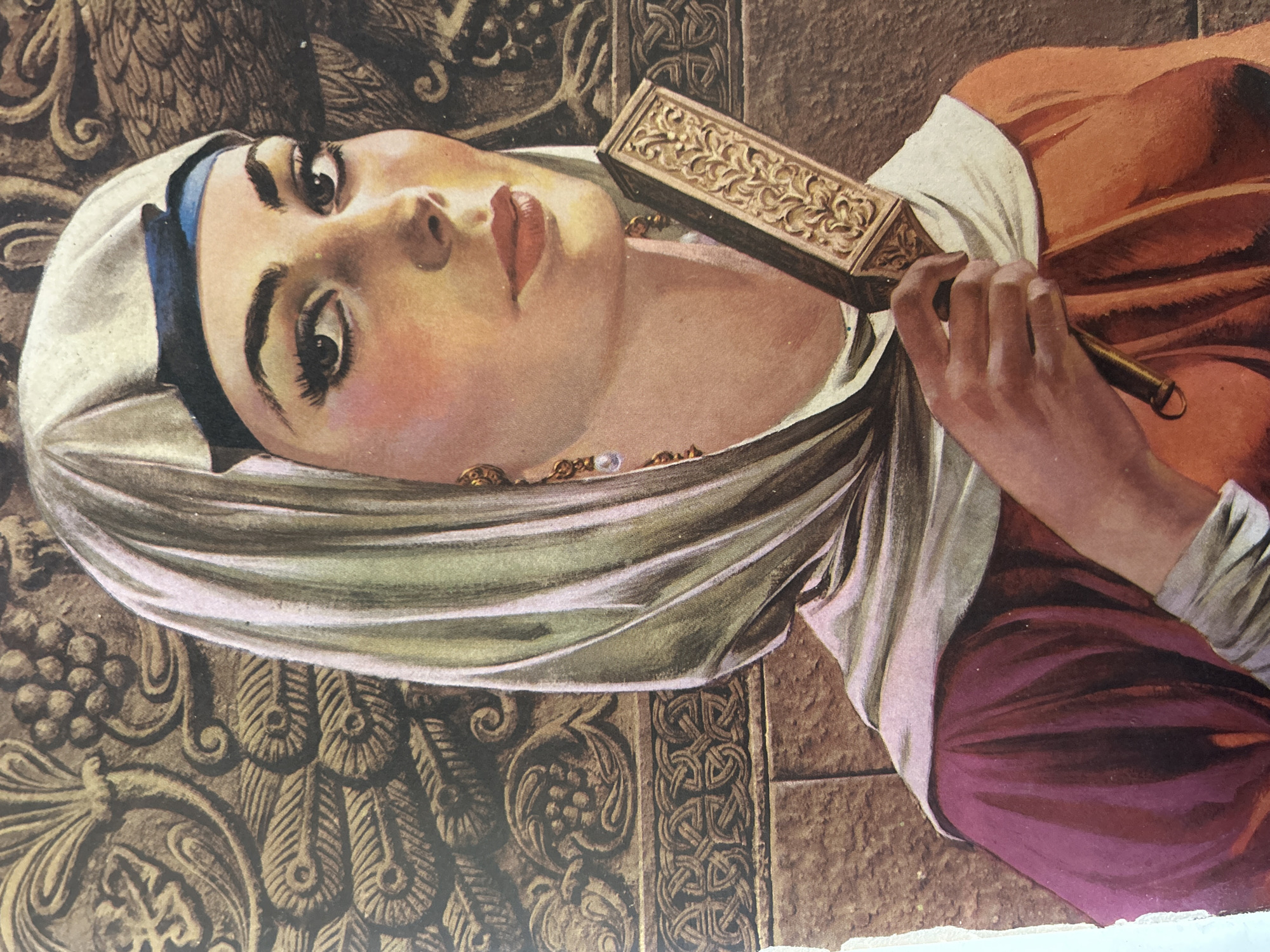 This is a close-up, painted portrait of a woman. She is looking to her right, towards the viewer, with her head slightly tilted. She wears a light-colored headscarf, possibly a hijab, draped over her head and shoulders, with a dark blue band visible beneath it. Her makeup is dramatic, with dark eyeliner and mascara, and rosy blush on her cheeks. She has red lipstick. She is wearing gold earrings, one of which has a pearl pendant.
The woman is holding a decorative, rectangular object in her right hand, which appears to be a handheld mirror with an ornate, gold-colored frame. Her left hand is not fully visible, but it seems to be positioned near her chest.
She is wearing a dark purple garment over an orange garment. The background behind her is an ornate, textured pattern in shades of brown and gold, resembling carved stone or tapestry. This decorative element features swirling motifs, leaf-like patterns, and a band of intricate knotwork.
There is no indication of Santa Ana, Venezuela, or any urban environment in this image. The setting appears to be an interior or stylized backdrop.