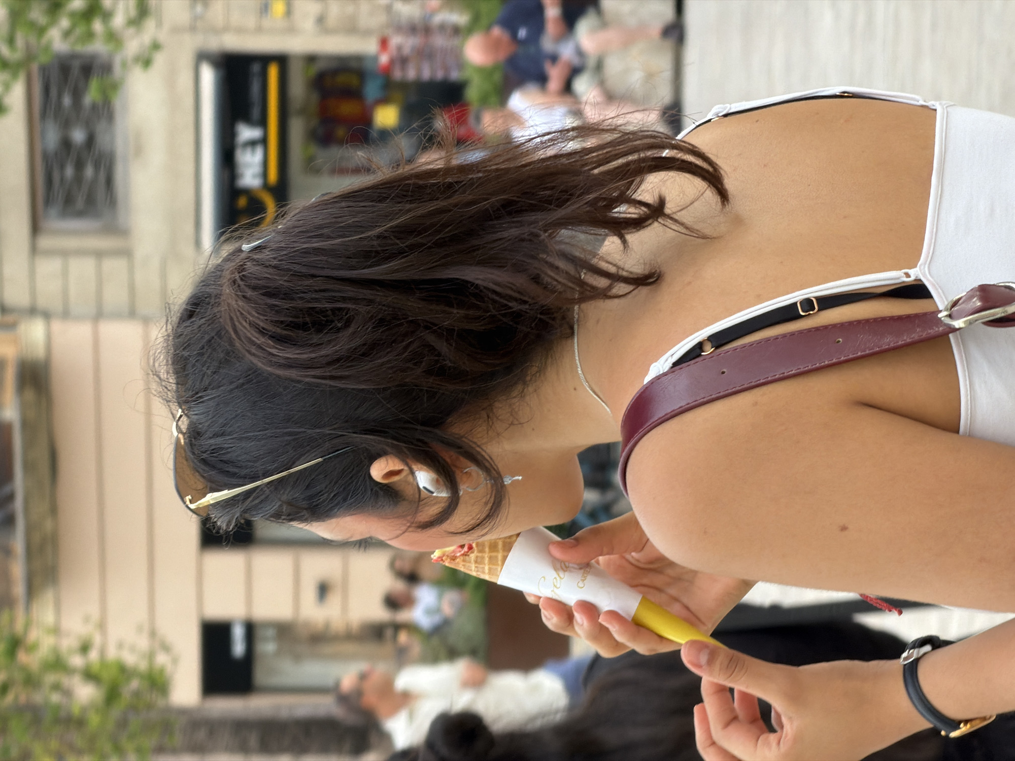 A woman is seen from behind eating an ice cream cone. She has dark hair pulled back in a ponytail, sunglasses on her head, and is wearing a white tank top with a burgundy strap. The woman is holding the ice cream cone, which is wrapped in white paper with gold lettering. Her left hand is visible, wearing a black watch with a gold face. In the background, a blurred scene of a city street with people and buildings is visible. The image is taken outdoors.