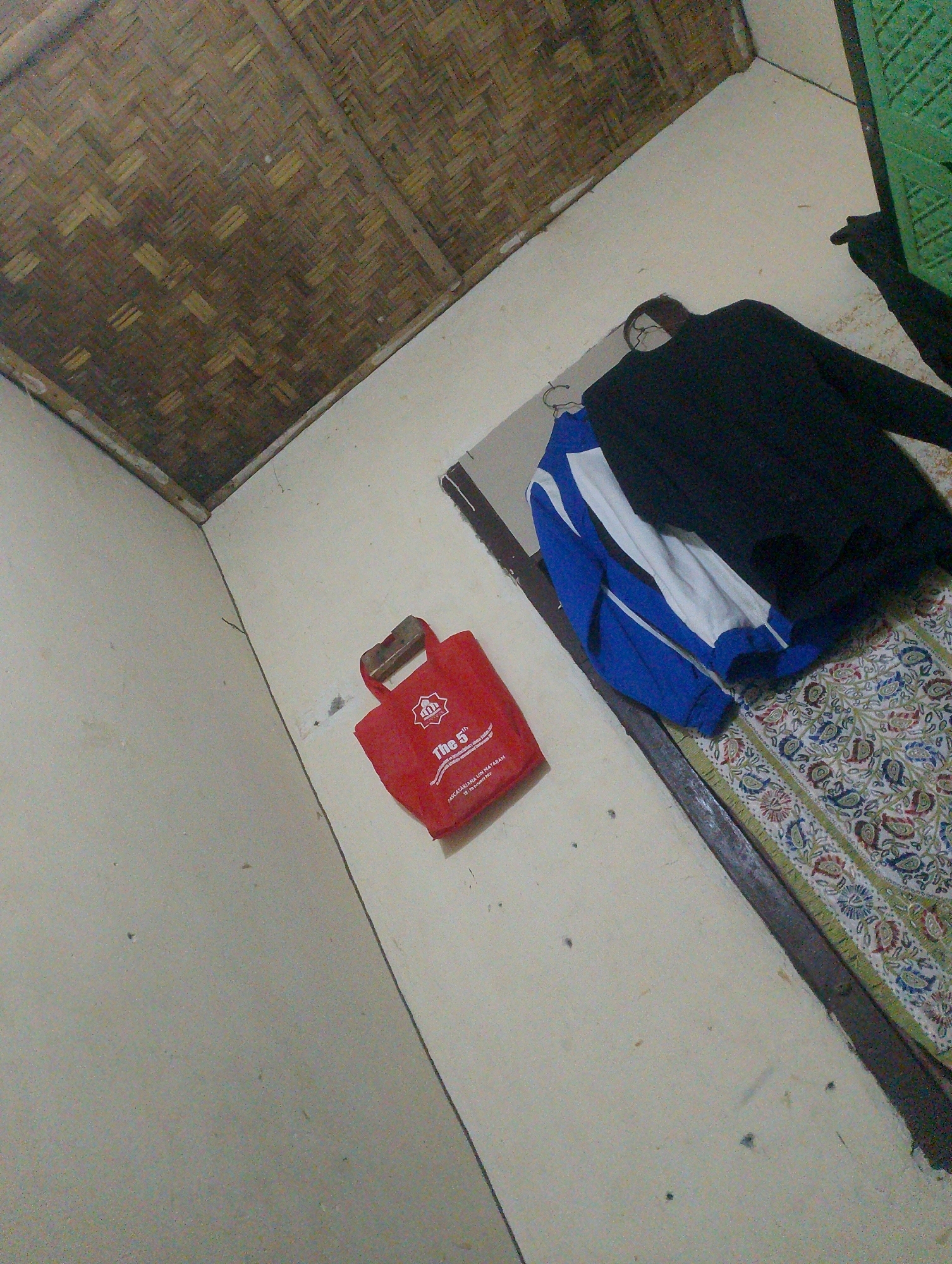 The image depicts a dimly lit interior room. In the foreground, the wall is a pale color. To the left, the ceiling is constructed of woven bamboo. In the middle ground, a red tote bag with white lettering that reads "The 5th" and an insignia is hung on the wall. Next to the bag, a blue and white jacket is draped over a black garment. The floor is covered with a rug featuring a floral pattern. The image appears to have been taken in Mujur, Indonesia.