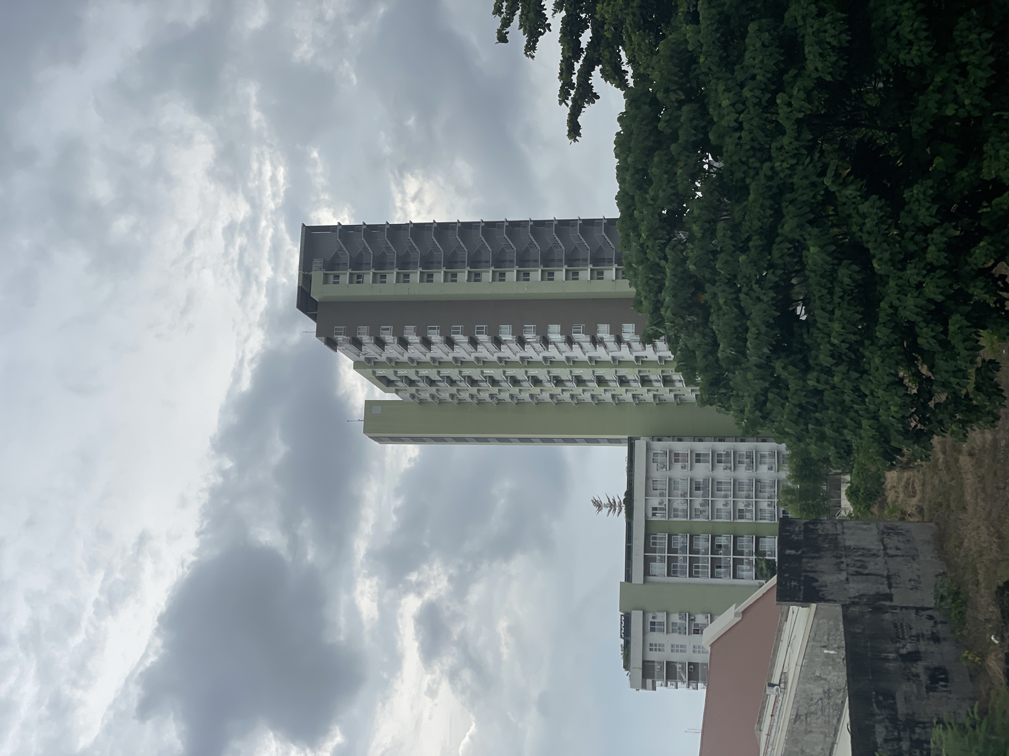 The image depicts an overcast day in Davao City, Philippines, with a view of several apartment buildings and lush green trees. In the foreground, a weathered concrete wall runs horizontally across the lower portion of the frame, with a reddish-brown roof of another structure visible to its left. Behind the wall, dry, sparse vegetation covers the ground.
Dominating the mid-ground are two apartment buildings. The building on the left is shorter and features a lighter green facade with multiple balconies. The taller building on the right has a darker green and brown color scheme, with visible external staircases running along its side. The apartment buildings appear to be modern high-rises.
In the background, dense green foliage from large trees partially obscures the sky. The sky itself is filled with heavy, grey clouds, suggesting impending rain or a generally cloudy day. The overall scene is a mix of urban architecture and natural elements, typical of a cityscape in a tropical region like Davao City. There are no people or discernible actions captured in the image.