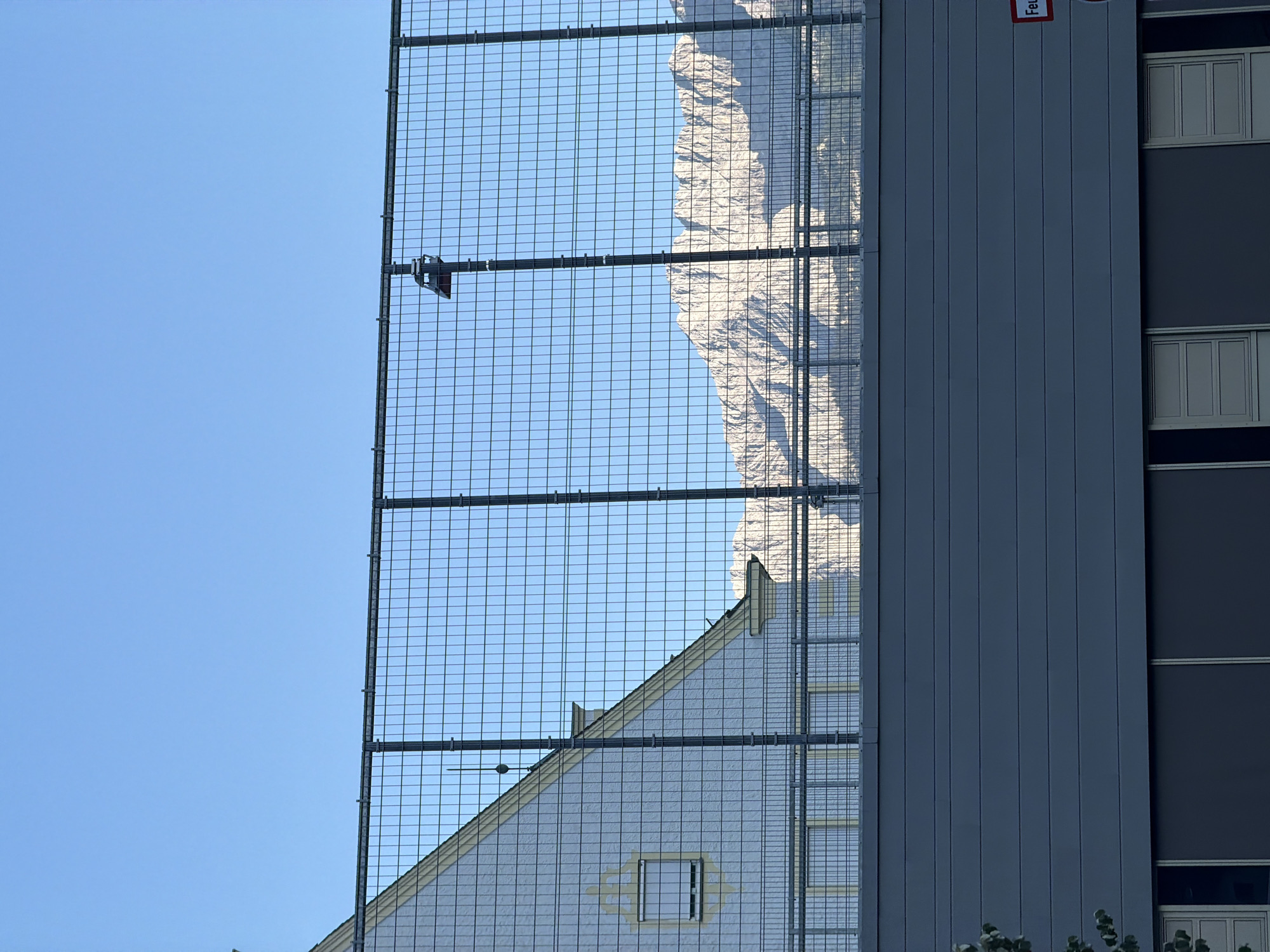 The image captures a view of the Zugspitze mountain range behind a building and a metal fence in Garmisch-Partenkirchen, Germany. The foreground features the grey, horizontally paneled facade of a modern building with several windows. Above this, a metal grid fence stretches across the frame, supported by vertical posts. Through the fence, the steeply sloped roof of a light-colored building is visible, with a single window. Further in the background, the jagged, snow-capped peaks of the Zugspitze mountains dominate the horizon under a clear blue sky. The scene is devoid of people.