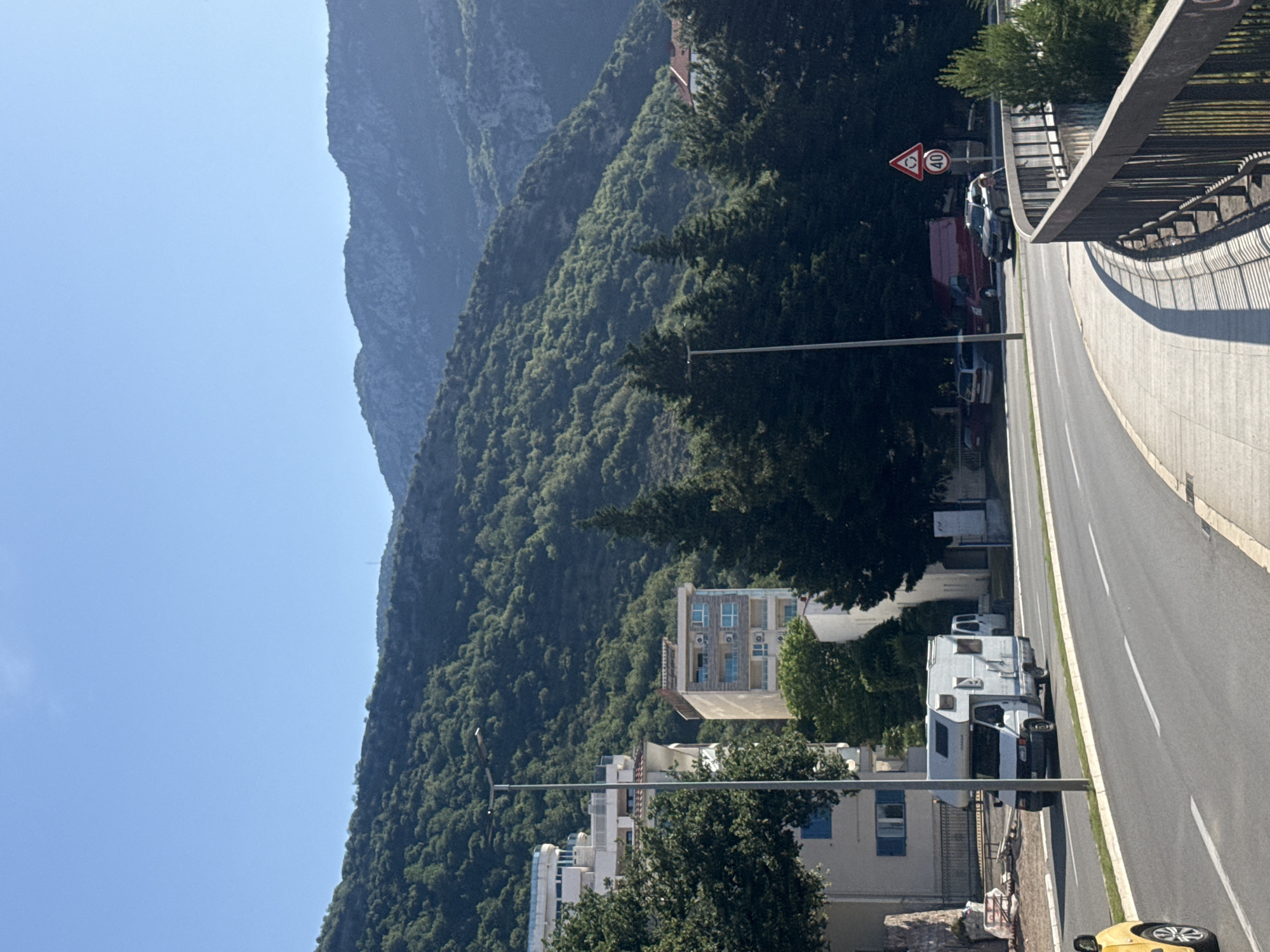 This image depicts a sunny outdoor scene in Rafailovići, Montenegro, with a focus on a multi-lane road curving through a populated area backed by forested mountains. In the foreground, an asphalt road curves to the right. It features a central median strip with a curb and sparse greenery, separating the visible lane from oncoming traffic. White double solid lines mark the center of the road, and a white solid line defines the right edge of the visible lane. On the far left, the partial front of a yellow car is visible. A white camper van with a raised roof is positioned on the road, traveling away from the viewer. To the right of the road, an elevated concrete sidewalk with a sturdy dark metal railing curves along the same trajectory as the road. Long shadows from the railing are cast onto the sidewalk. Further down the road on the right, several vehicles are parked: a grey SUV, a red cargo van, and a silver sedan. A person on a bicycle is partially visible behind the grey SUV. Two road signs are visible on the right: a red triangular warning sign with a black car symbol (likely indicating road narrowing or a hazardous curve) and a circular white sign with a red border indicating a "40" speed limit. Tall, dark green coniferous trees are interspersed along the roadside and behind the railing on the right. On the left side of the road, beyond the median, several multi-story buildings are visible. One prominent white building features multiple balconies and a distinctive curved upper floor. Further back, another building displays light grey/beige facades with blue-tinted balcony railings and several external air conditioning units. A white building with blue window shutters is also visible closer to the foreground. A modern streetlight with a flat LED fixture stands centrally on the left side of the road. The background is dominated by a succession of mountains. The lower and middle slopes are densely covered with lush green foliage, suggesting extensive tree growth. Higher peaks, further in the distance, appear rockier and less vegetated, displaying greyish hues under the bright sunlight. The sky above is clear and bright blue, with a very faint wispy cloud formation visible near the top left. Another streetlight is visible in the midground on the right, mirroring the one on the left. A small white building with a terracotta-colored roof is partially
