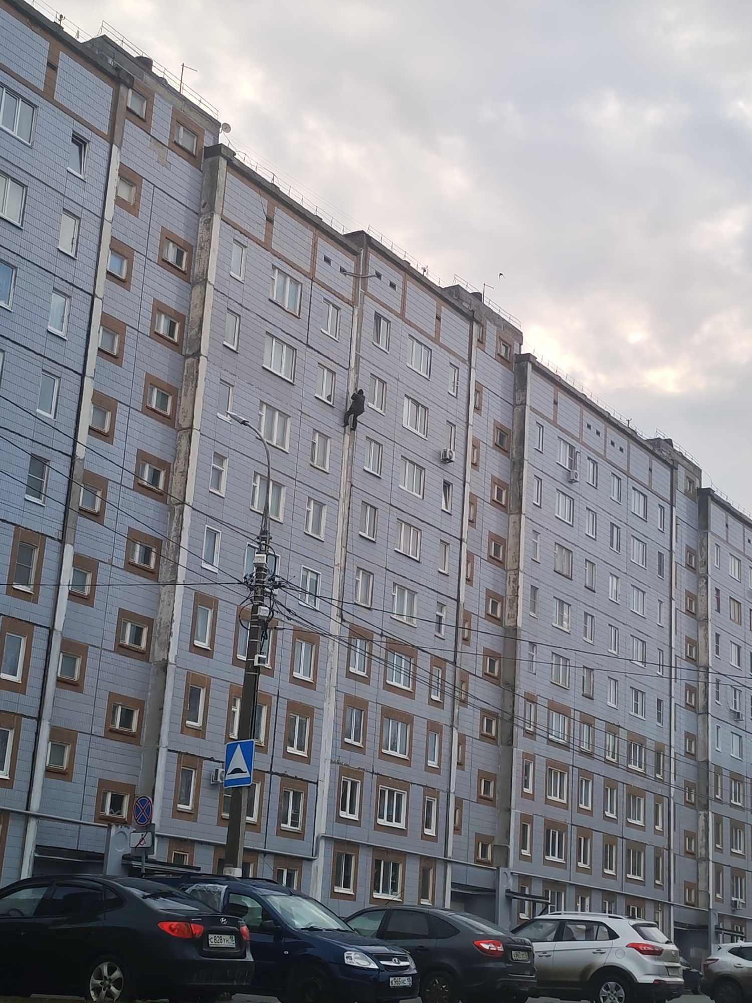 The image shows a gray apartment building in Izhevsk, Russia, with multiple windows. A person is scaling the side of the building via a vertical structural element. In the foreground, parked cars line the street. Overhead, a cloudy sky dominates the scene. A utility pole with wires and a street sign is visible in front of the building.