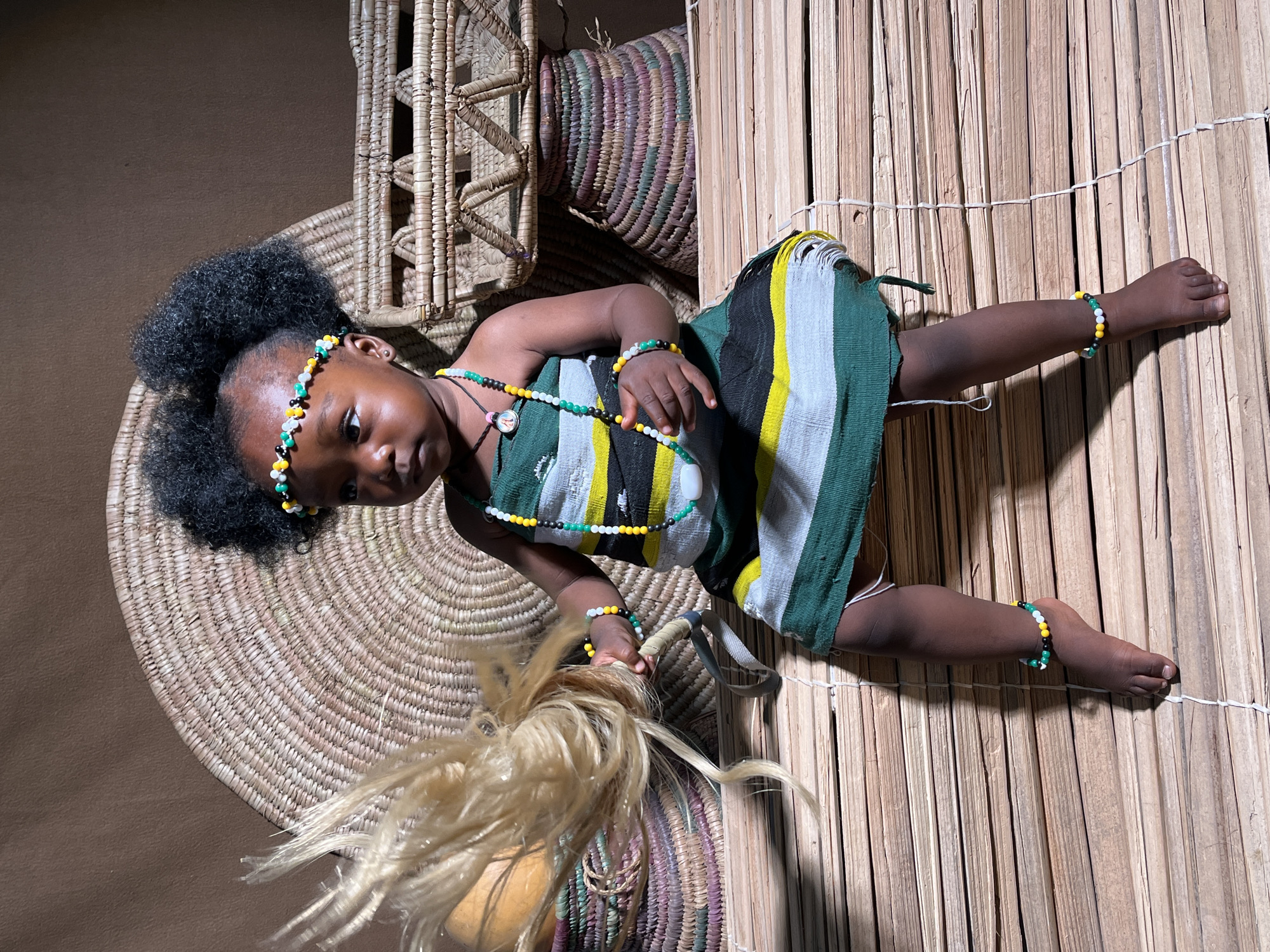 A young child is lying on a reed mat in a studio setting, with woven baskets and a straw fan placed around. The child is dark-skinned, with afro-textured hair adorned with a beaded headband. They are wearing a striped green, yellow, black, and white dress, and multiple beaded jewelry pieces on their head, neck, wrists, and ankles. The child's gaze is directed toward the camera. The background includes various woven items and a dark, unidentifiable surface. The location of Barakin Akawo, Nigeria, is not discernible from the image content alone.