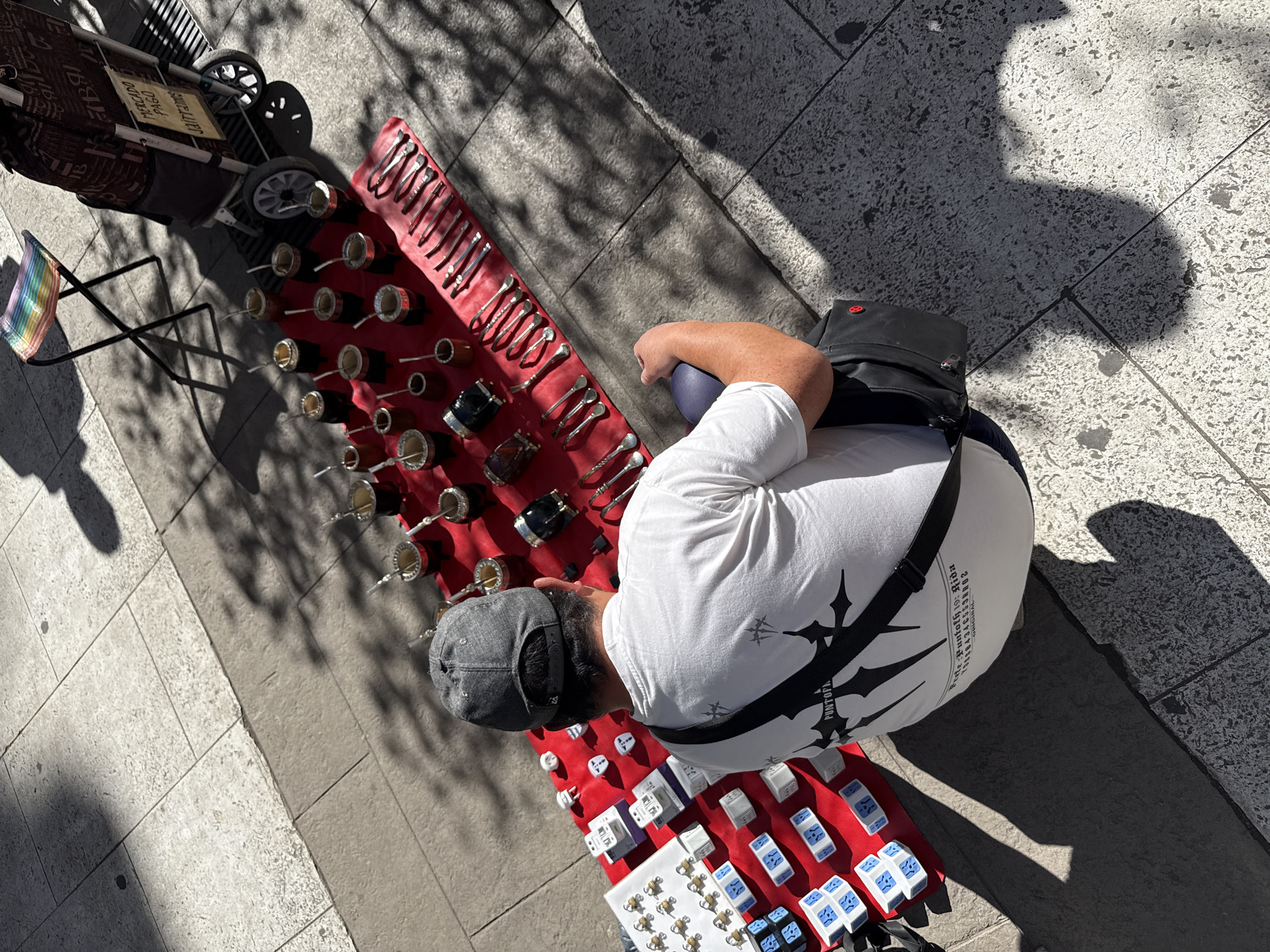 The image is an overhead view of a street vendor in Buenos Aires, Argentina, displaying merchandise on red cloths laid out on the pavement. The vendor, wearing a grey cap, white t-shirt, and a black messenger bag, stands near their display. The merchandise includes mate cups with bombillas, various spoons and tongs, rings and other small items. A shopping cart with a sign and a small rainbow-colored folding chair are positioned next to the display. The scene is bathed in sunlight, creating shadows from the trees and surrounding objects. The pavement consists of large, light-colored tiles.