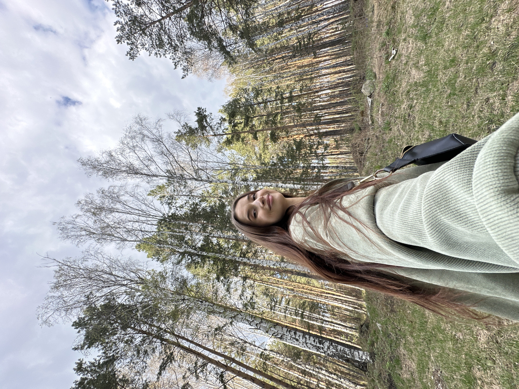 A woman poses for a selfie in a forest in Yekaterinburg, Russia. The woman has long brown hair and is wearing a light green jacket. She is standing on a grassy area next to a forest of birch and pine trees. A black bag is visible. The sky is cloudy.