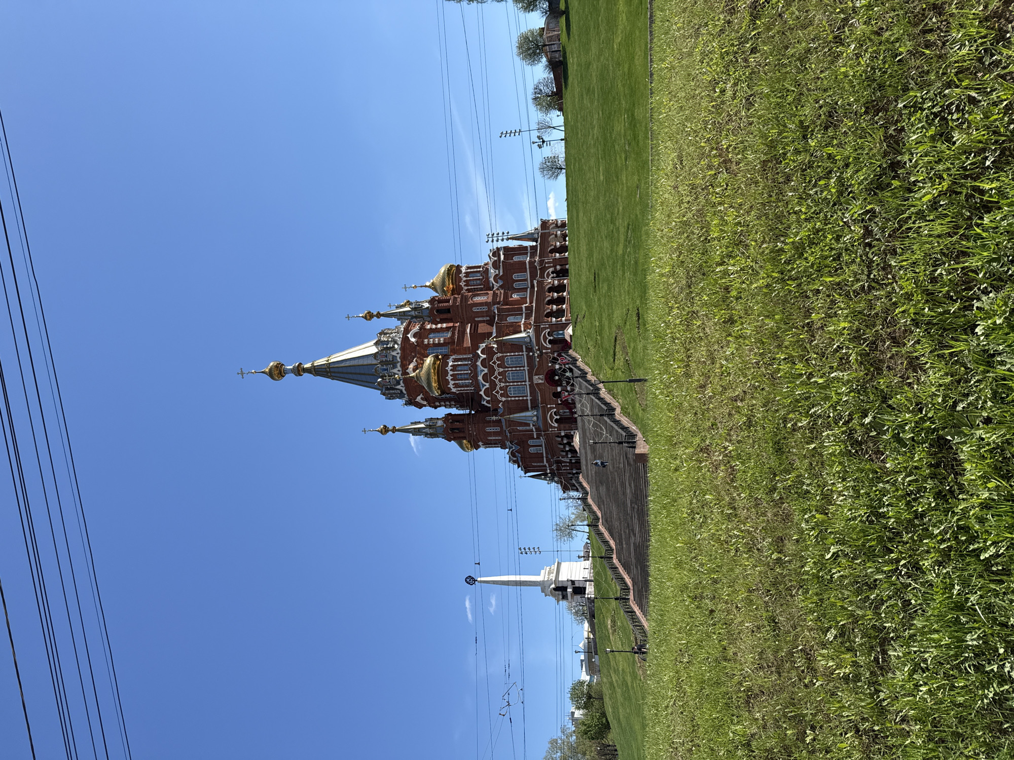 The image shows the Saint Michael's Cathedral in Izhevsk, Russia, taken from a low angle. The cathedral is brick red with gold domes and a tall spire. A white obelisk stands nearby. Lush green grass covers the foreground and background. Overhead power lines are visible against a clear blue sky. There are no people visible.