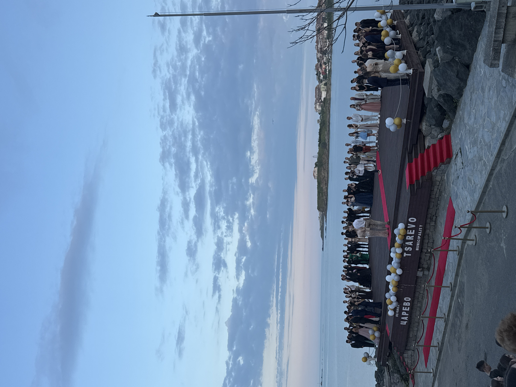 The image depicts a gathering of people on a wooden platform or stage adorned with a red carpet and white and gold balloons. "TSAREVO MUNICIPALITY" is written in both Bulgarian and English on the stage. The stage is situated near a rocky cliff overlooking the sea. In the background, a coastline with buildings is visible. The sky is overcast with clouds. It is likely a celebratory event or graduation ceremony taking place in Tsarevo, Bulgaria.