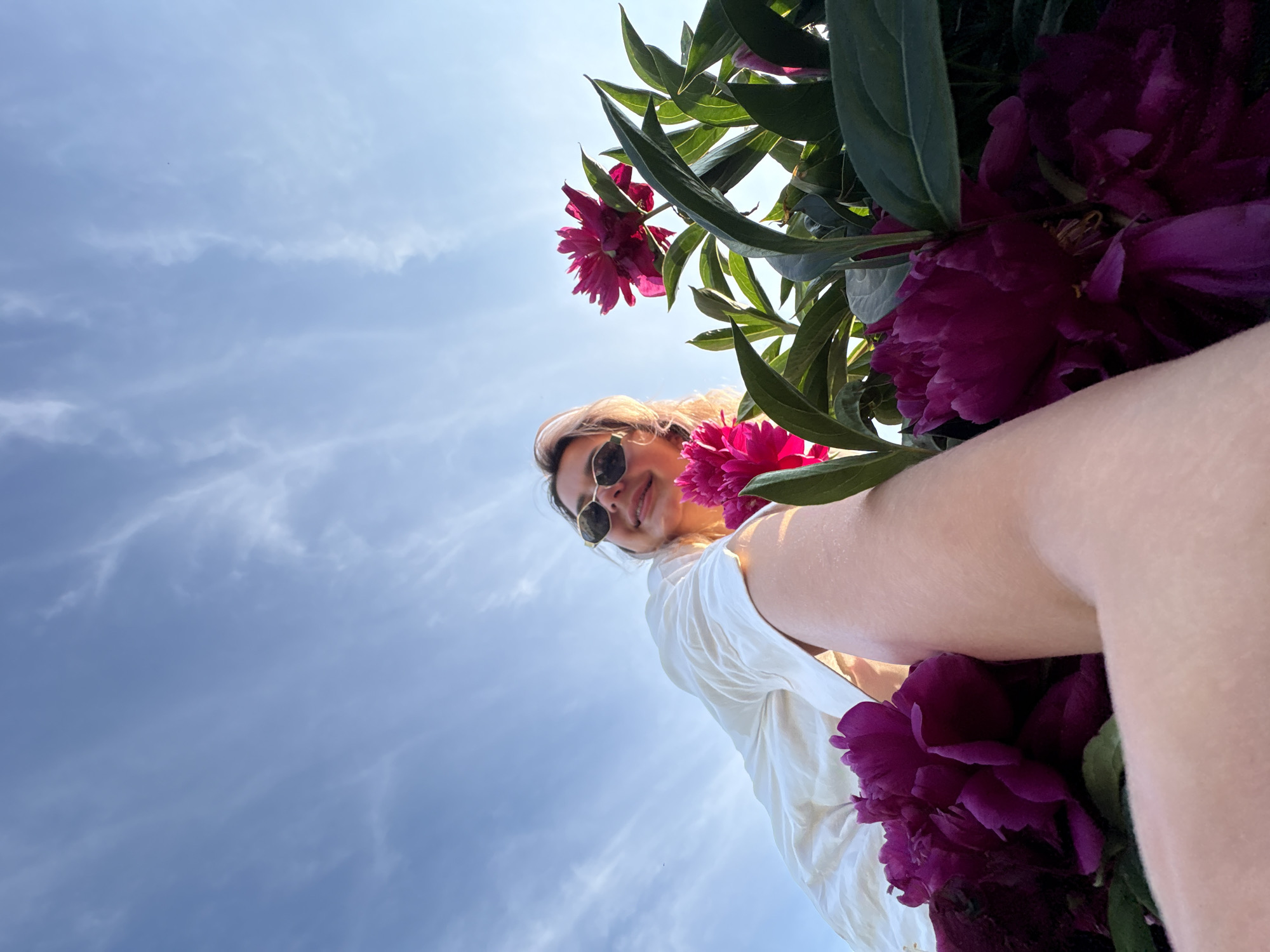 A low-angle, close-up shot captures a woman from the chest up, partially obscured by vibrant pink peonies. She is wearing a white collared shirt and sunglasses with geometric frames. Her blonde hair is visible, and she is smiling. The background is a bright blue sky with wispy white clouds. The scene is outdoors, and the presence of peonies suggests a garden or park setting. The image was taken in Razdolnoye, Russia.