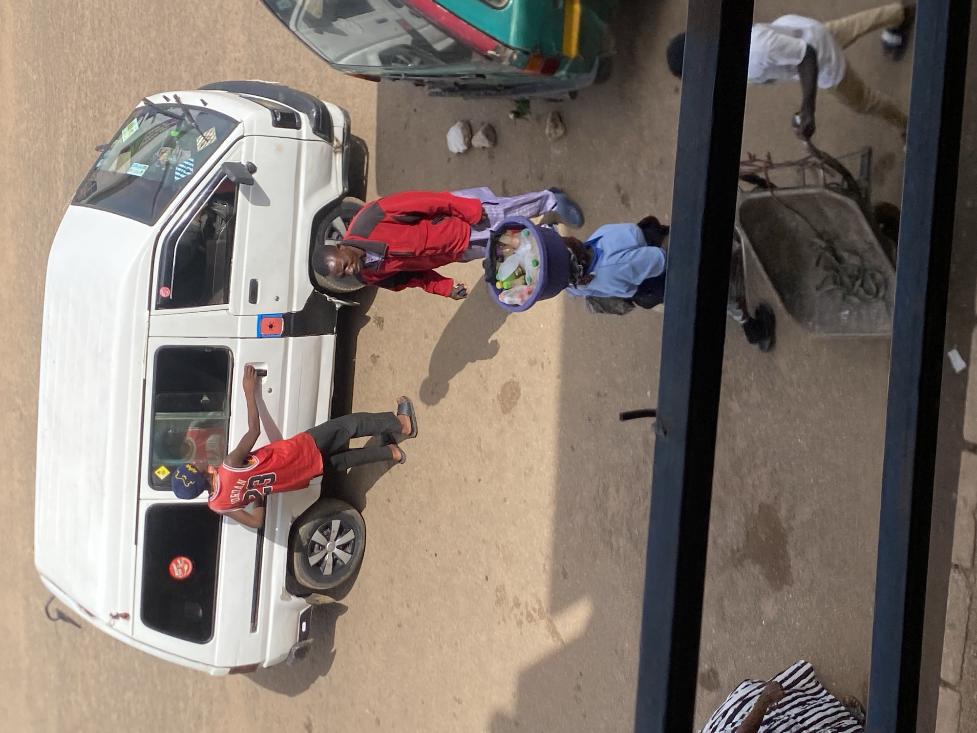 This media file depicts an outdoor scene in Bukuru, Nigeria, likely during daylight hours as indicated by the presence of shadows. The primary subjects are several individuals and a white van. One person, wearing a red jacket and light-colored pants, stands next to the van, looking towards the viewer. Another person, dressed in a blue shirt and light pants, is crouched down near a large blue basket filled with various bottles, possibly beverages. A third individual, wearing a red basketball jersey with the number '23' and a blue baseball cap, is leaning against the side of the white van, with their arm on the door handle. In the background, a green vehicle is partially visible, and a person in a white shirt and tan pants is seen operating a wheelbarrow. The ground appears to be a dusty or paved surface. The scene suggests daily life activities, perhaps related to commerce or transportation. The overall mood is neutral, with no strong emotional cues evident from the subjects' expressions or postures. The image is captured from an elevated viewpoint, looking down at the scene.