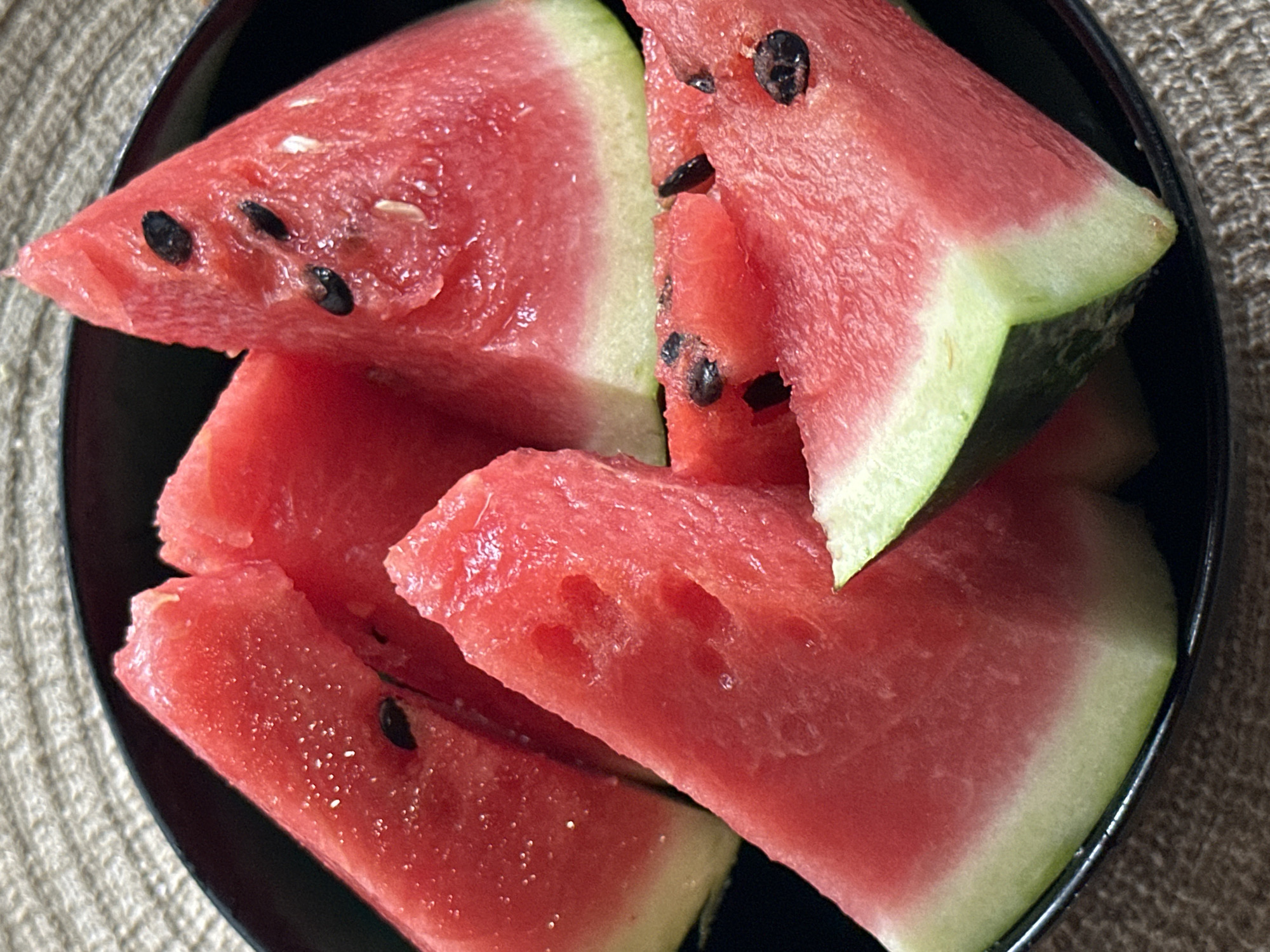 A black bowl filled with watermelon slices is displayed on a woven mat. The watermelon slices are triangular and juicy, with some showing visible black seeds. The flesh of the watermelon is a vibrant red, transitioning to a pale green rind at the bottom. The lighting is bright, highlighting the texture and moisture of the fruit. The scene is a close-up shot, focusing on the details of the watermelon and the bowl.