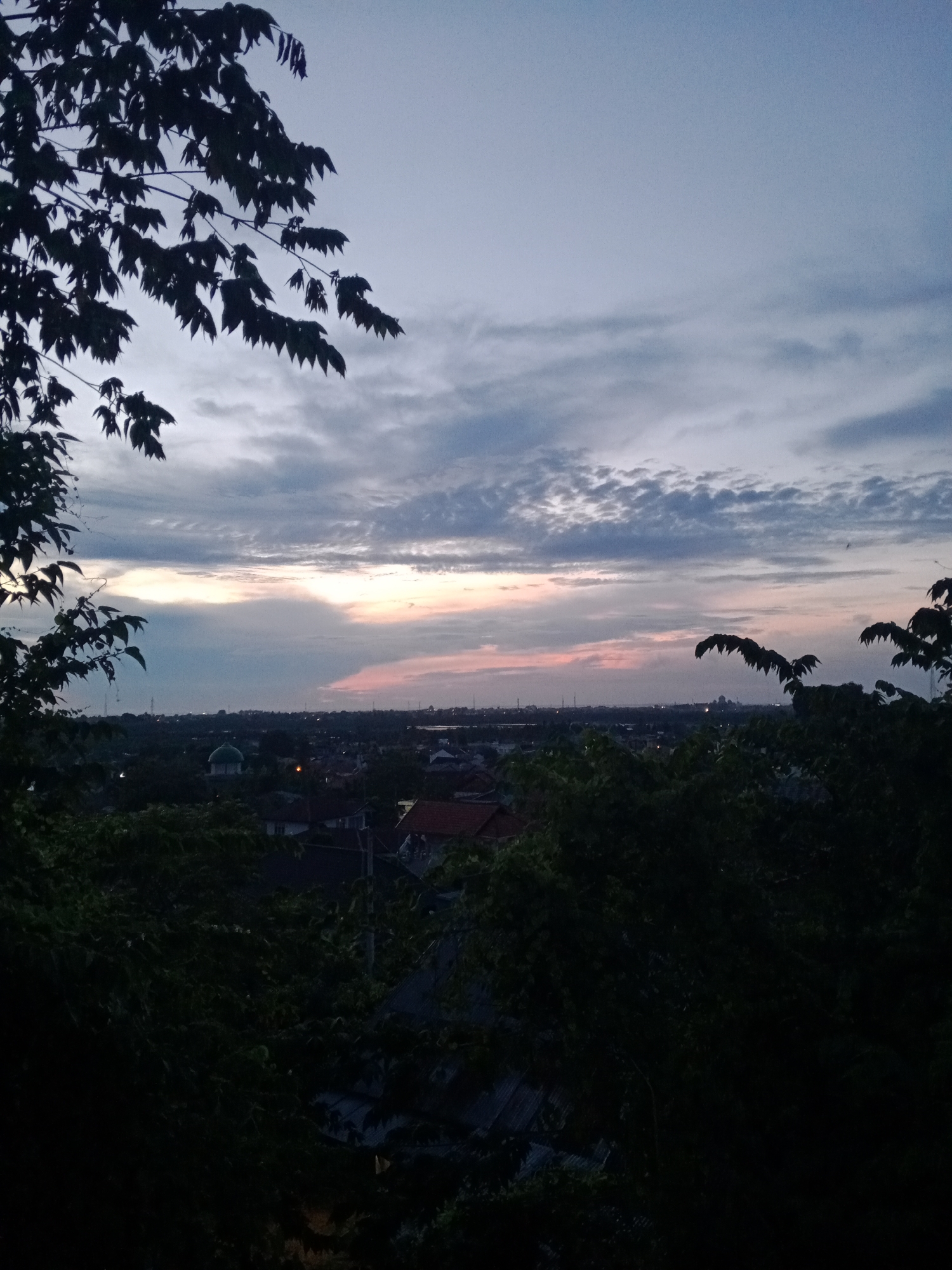 The image shows a dusk or dawn scene in Lhokseumawe, Indonesia. In the foreground, dark silhouettes of trees frame the view. Beyond the trees, the cityscape is visible, with various buildings and houses with rooftops. A prominent green-domed building, likely a mosque, stands out among the structures. In the distance, a faint, reddish-orange glow suggests the horizon line where the sky meets the land or sea. The sky is filled with clouds, transitioning from darker shades above to lighter hues near the horizon. Overall, the scene captures an urban landscape in Indonesia during a transitional time of day.