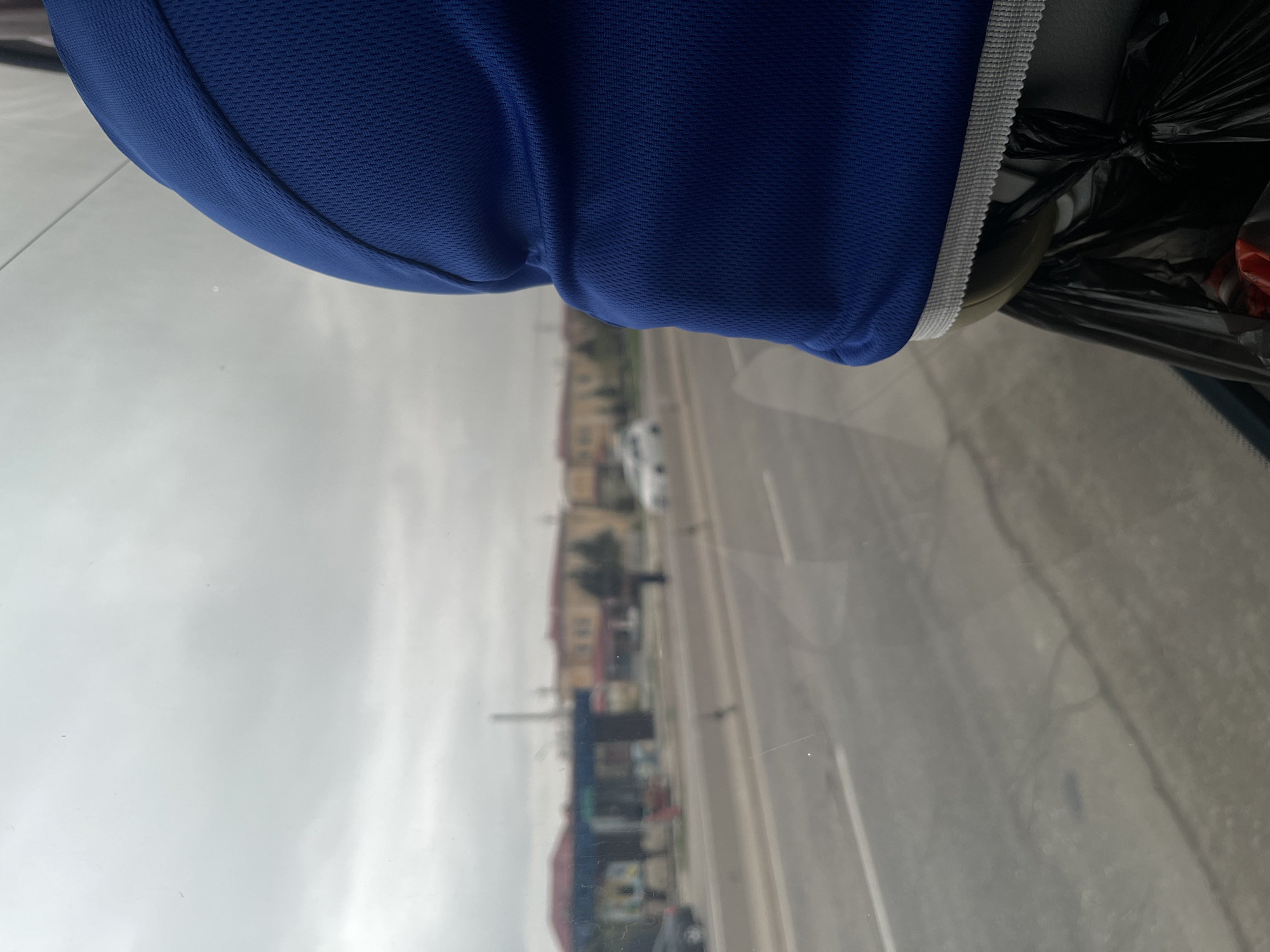 The view is from inside a vehicle, looking out onto a street with buildings on either side. The foreground includes a blue athletic garment with a white trim, partially obscuring the view. A black plastic bag is also visible. The street has multiple lanes of traffic with vehicles visible. The buildings along the street appear to be a mix of residential and commercial properties, possibly a shopping area. The sky is overcast. While the location is purported to be Karmana, Uzbekistan, there is no definitive evidence within the image to confirm this claim.
