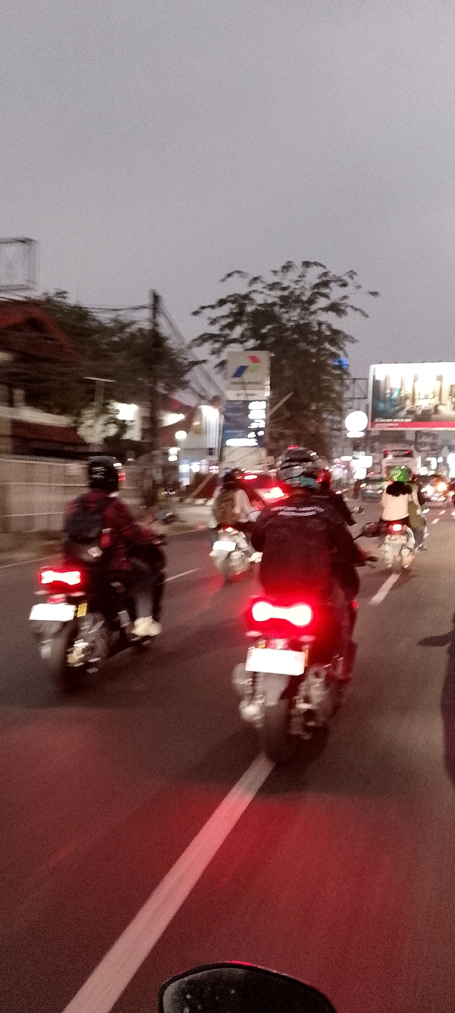 The image depicts a street scene in Bekasi, Indonesia, likely during dusk or early evening, given the ambient light and illuminated vehicle lights. Several motorcycles are visible, some with riders, moving along a paved road. The foreground shows the rear of a motorcycle with its distinctive heart-shaped red taillights illuminated. Another motorcycle with similar taillights is further ahead. In the middle ground, more motorcycles and riders are present, along with the blurry outlines of buildings and trees on the left side of the road. A prominent sign with a colorful logo, possibly advertising a fuel company, is visible in the background, along with other illuminated signage. The overall impression is of moderate traffic flow on a city street.