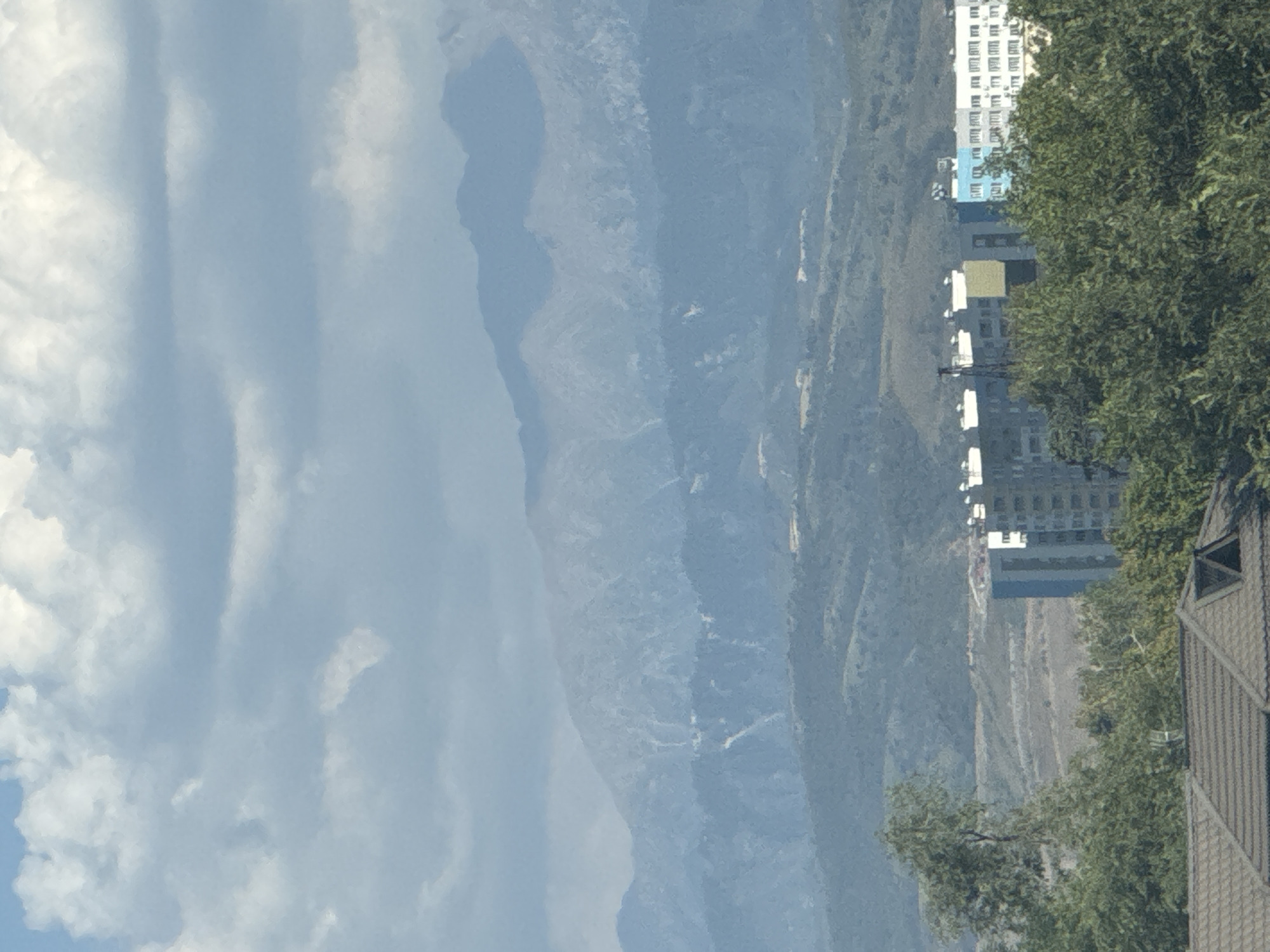The image shows a mountainous landscape behind a city skyline. In the foreground, there are trees and the roofs of buildings. Further back, there are several multi-story buildings, some of which are colored blue and white, with a yellow rectangular section on one building. Behind the city, rolling hills are visible, and beyond those, progressively more distant and hazy mountain ranges. The sky is partly cloudy, with large, fluffy white clouds against a pale blue sky. This view is characteristic of Almaty, Kazakhstan, which is known for its proximity to the Tien Shan mountains.