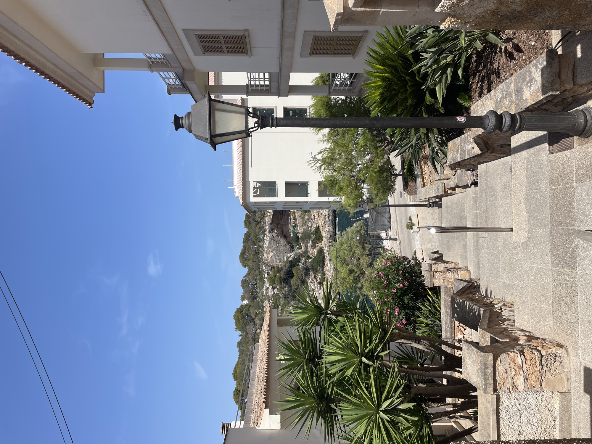 This image shows a view looking down a set of stone steps in Cala Figuera, Spain. The steps are flanked by stone walls and lush green vegetation, including large yucca plants. A black lamppost stands prominently on the right side of the steps, casting a shadow. In the background, white buildings with balconies and brown-tiled roofs are visible, as well as the blue sea and rocky cliffs covered with trees. The sky is clear blue with a few wispy clouds. There are no people or animals in the frame.
