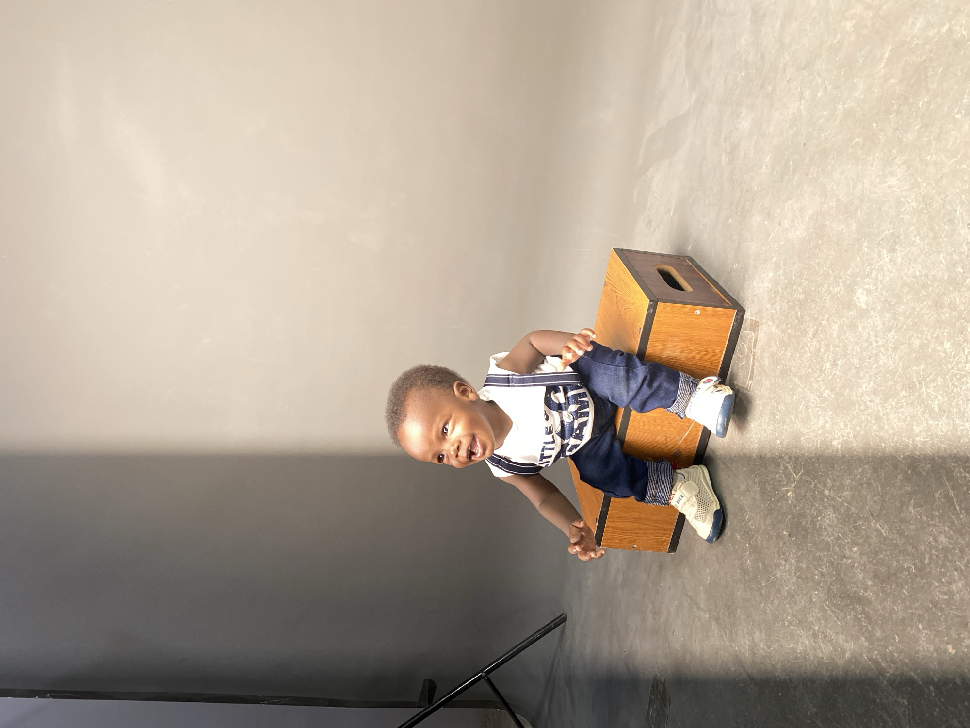 A baby sits on a wooden box in a room with a gray floor and wall. The baby wears a white shirt with blue accents and dark blue pants, along with white and blue shoes. The baby is smiling and facing the viewer. A portion of what appears to be a light stand is visible in the lower-left corner. The location is Gura Top, Nigeria.