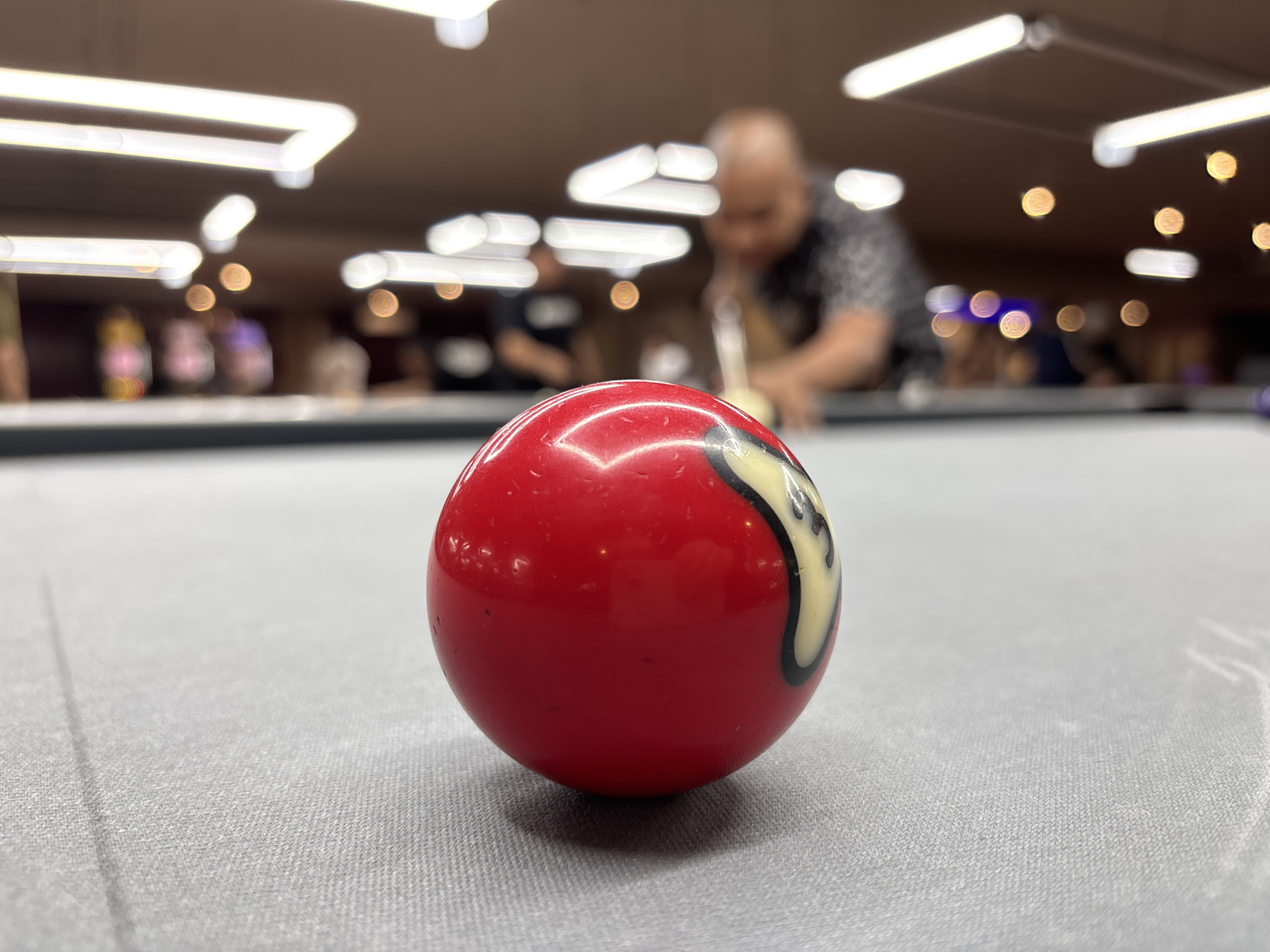 A close-up, low-angle shot focuses on the 8-ball of a pool table, set against a backdrop of a dimly lit billiard hall in Kuta Selatan, Indonesia. The 8-ball, with its prominent black number 8 on a white circle, is positioned slightly left of center, its polished red surface reflecting the overhead lights. The pool table's felt surface is a muted gray. In the background, out of focus, a person is seen leaning over the table, preparing to take a shot with a cue stick. Other blurred figures and patrons are visible further back, adding to the ambiance of a recreational establishment. The environment appears to be an indoor entertainment venue, characterized by a warm, ambient glow from the ceiling lights and some decorative lighting in the distance.