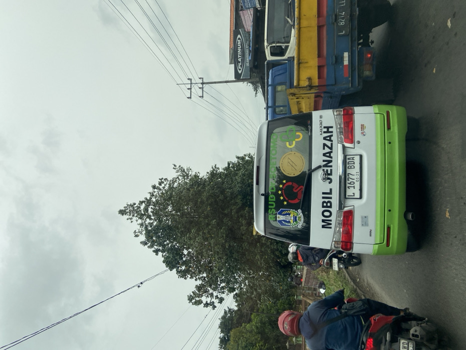 A white and green "Mobil Jenazah" (Funeral Car) with license plate L 1677 BDA is positioned on a road. Stickers on its back include "RSUD Dr. Soetomo" and other logos. A motorcycle is right behind the car. A large truck is visible alongside the car, next to a "Platinum" sign. Overhead, power lines traverse the scene against a cloudy sky.