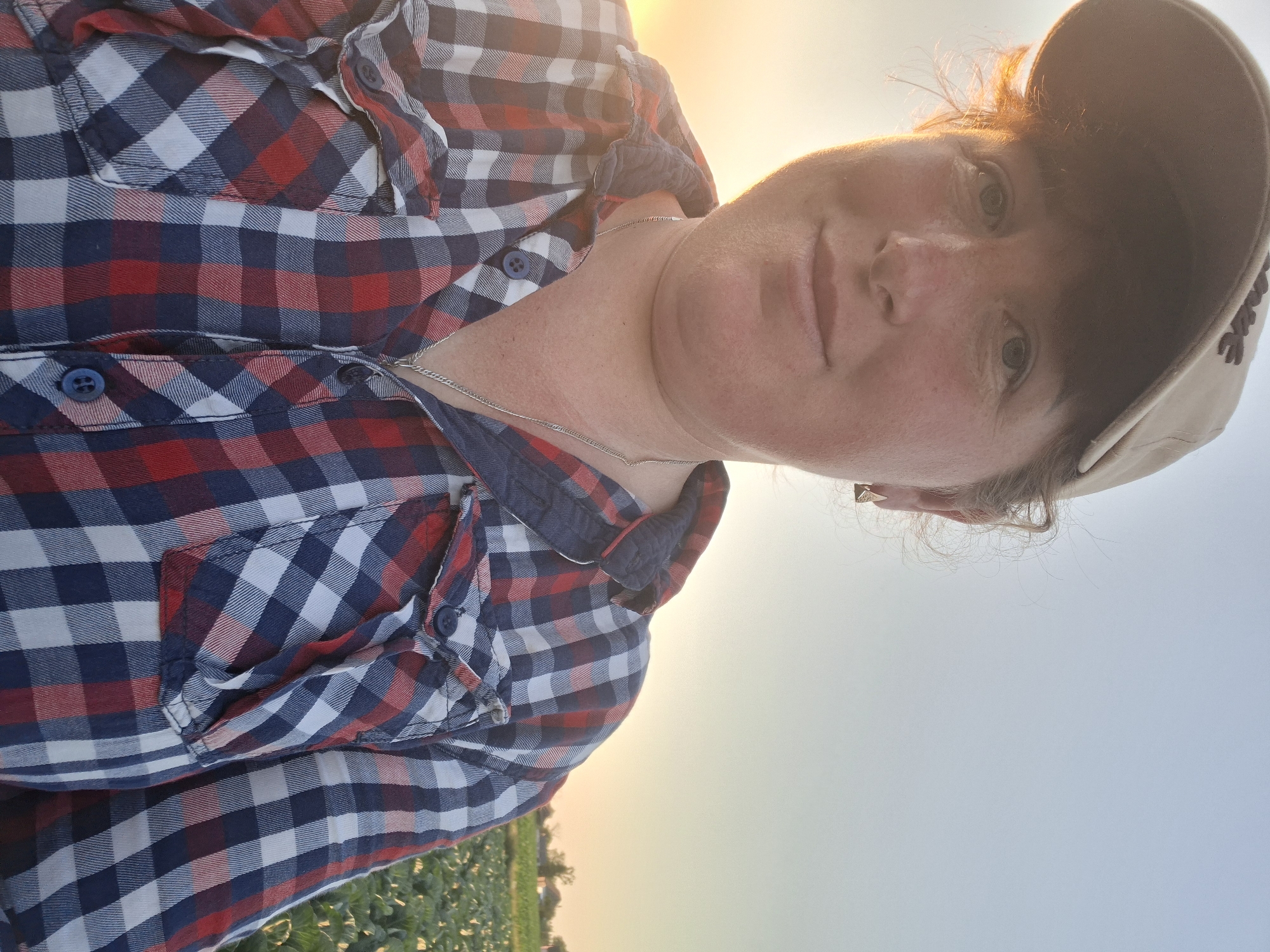This is a portrait-style photograph of a person wearing a red, white, and blue plaid button-down shirt and a tan baseball cap. The image is oriented sideways, with the person's head towards the top right and their body extending towards the bottom left. The person is looking at the camera with a slight smile. They have fair skin, freckles, and light brown hair that is visible under the brim of the cap. A silver necklace is visible around their neck.
The background is a hazy sky, suggesting either sunrise or sunset. The warm glow on the person's face and hair indicates this as well. In the bottom left corner of the image, there is a hint of a green field, possibly crops, with some trees in the distance. The lighting suggests it is late afternoon or early evening. The overall mood of the image is relaxed and natural. There is no visible text in the image.