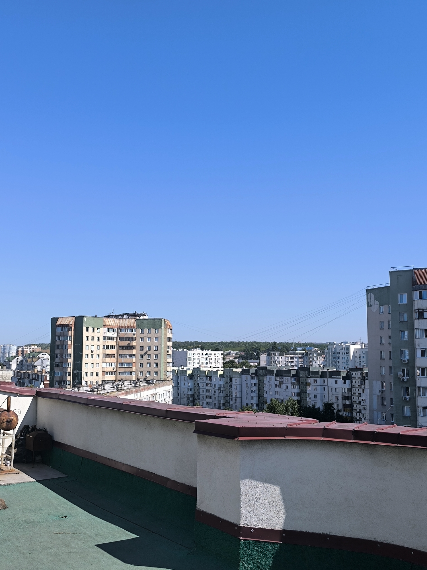 This is a high-angle shot of a cityscape on a clear, sunny day, likely taken from a rooftop. The foreground features a rooftop with a green surface and a white parapet wall topped with a brown roof trim. To the left of the parapet, there are some grills and what appears to be a small metal structure. In the mid-ground and background, a dense arrangement of Soviet-era apartment blocks fills the horizon. The buildings are of varying heights, with many exhibiting a uniform, blocky design characteristic of this architectural style. Colors range from light gray and white to muted greens and browns on the building facades. Some buildings have visible balconies and window units. A thin, dark line of trees is visible in the distance, separating the apartment blocks from the sky. The sky itself is a vast expanse of clear, bright blue. Based on the architectural style of the buildings, the scene is likely located in Chișinău, Moldova.