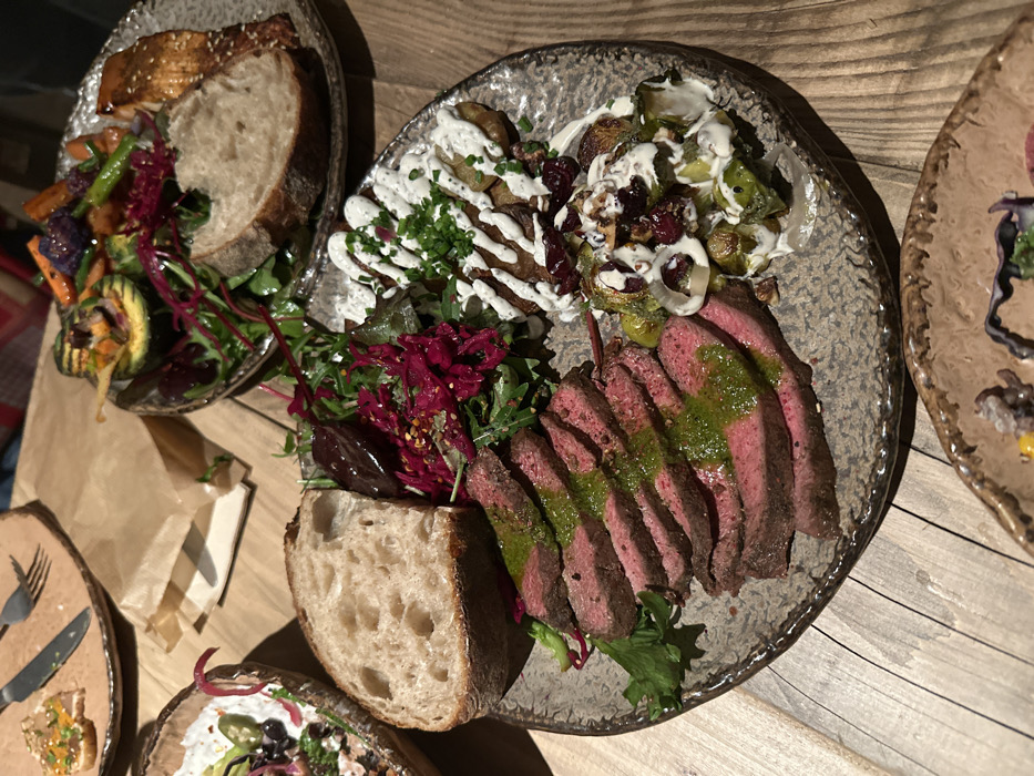 Several plates of food are arranged on a wooden surface. One plate holds sliced steak drizzled with green sauce, a slice of bread, and a side salad with red leaves. Another plate features cooked potatoes and Brussels sprouts with a white drizzle and cranberries. A third plate contains grilled salmon, vegetables, and bread. Cutlery and a paper bag are also visible on the table.