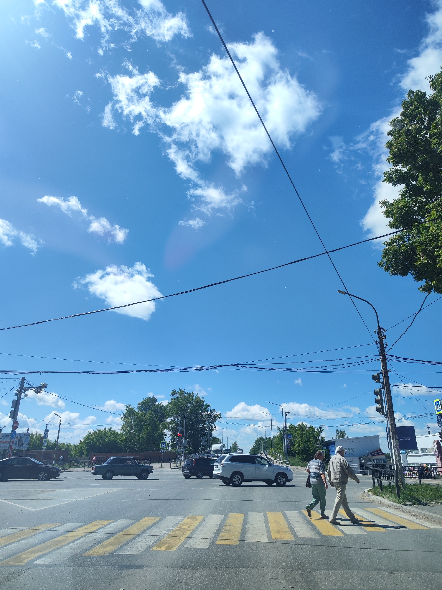 The image captures a street scene in Chaikovsky, Russia, with a bright blue sky and scattered white clouds overhead. Multiple power lines crisscross the frame, supported by utility poles. A green tree on the right side of the image extends into the frame from the top. In the foreground, a pedestrian crossing is marked with yellow and white stripes on the asphalt. Two people are walking across this crossing from left to right. The man on the right is wearing a light-colored jacket and pants, while the person on the left is in a striped shirt and green pants. Several cars are visible on the road, including a dark sedan, a dark SUV, and a white SUV. Traffic lights and street signs are present at an intersection. Buildings, including one with a sign that reads "ОРИОНА" (ORION) in reverse, are visible in the background. Trees and foliage line the sides of the street.