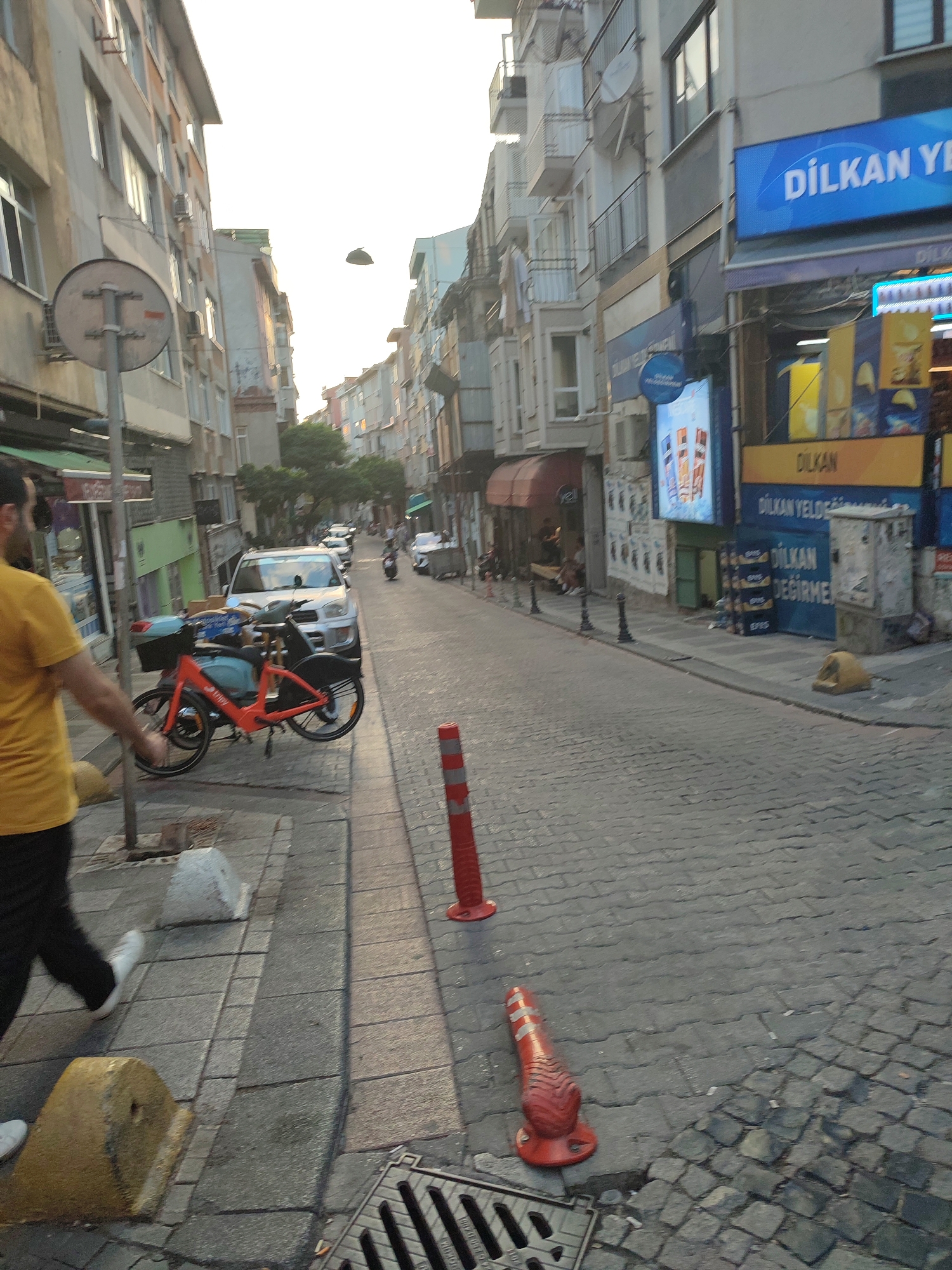 The image shows a street scene in Kadıköy, Turkey, on a paved road lined with buildings. A person wearing a yellow shirt and black pants is walking on the sidewalk. An orange bicycle is parked on the sidewalk. A white SUV is parked on the street. Several shops are visible, including one with a blue sign that says "DILKAN YEN". Traffic cones are placed on the road. The sky is overcast, suggesting it may be early morning or late afternoon. The overall impression is that of a typical urban street in Turkey.