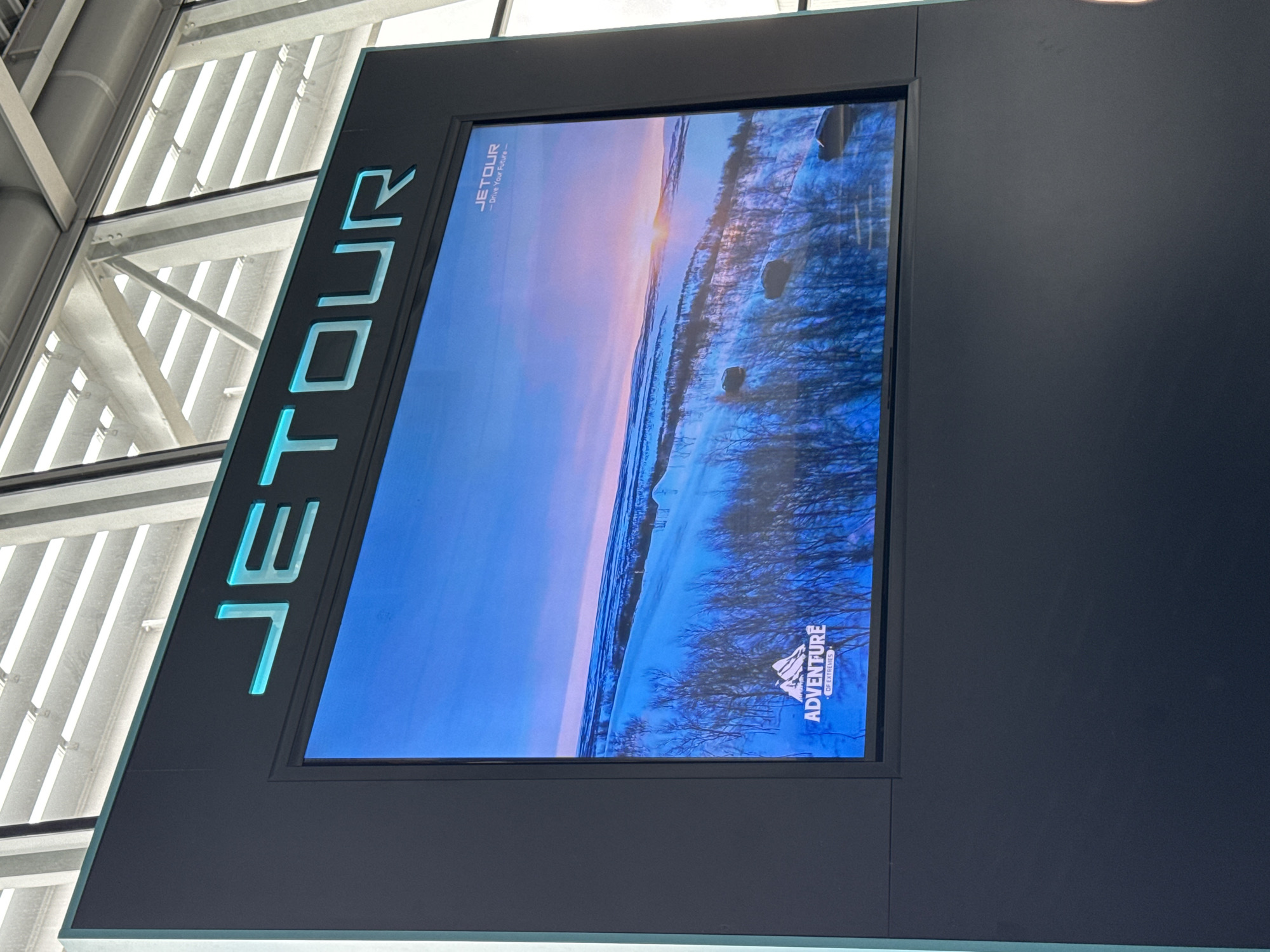 The image displays a large, vertically oriented digital screen advertising "JETOUR" within a modern indoor setting, likely in Warsaw, Poland. The screen features a black frame with the brand name "JETOUR" prominently displayed in large, light blue, stylized letters running vertically along the left edge.
The content on the screen shows a serene winter landscape, suggesting either sunrise or sunset. The sky transitions from soft pink and orange hues near the horizon, where a bright light source (presumably the sun) is visible, to a pale blue higher up. The ground is covered in snow, with numerous bare trees or bushes with dark branches, indicative of a cold climate.
Within the digital advertisement, smaller white text reading "JETOUR" and the tagline "-Drive Your Future-" appears in the top-left corner. In the bottom-right corner, a logo featuring a mountain icon is present, along with the text "ADVENTURE" and "OF EXTREMES," reinforcing themes of travel and exploration.
The background reveals modern architectural elements, including glass panels and white metal support structures, typical of an airport or a large public building, where such a display would be common for promotional purposes.
