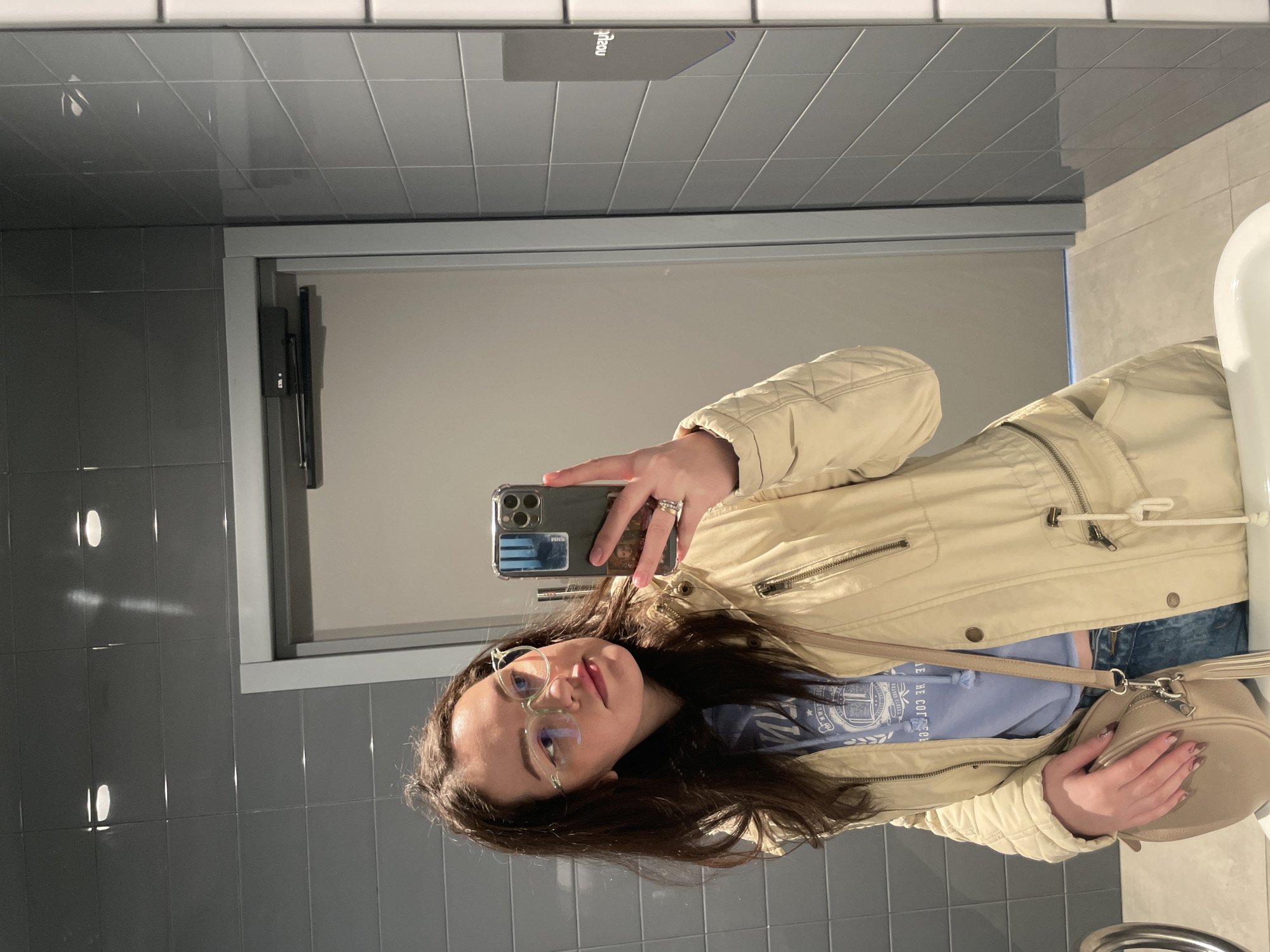A young woman is depicted taking a mirror selfie in what appears to be a public restroom. She is wearing a light beige or cream-colored quilted jacket over a light blue hoodie or long-sleeved shirt with some white text, and blue jeans are partially visible. She has long, dark hair, clear-framed glasses, and a light beige shoulder bag slung across her body. A ring is visible on her left hand, which holds a smartphone with a clear case that contains visible elements like "SWIM" and "HM" stickers or cards.
The background consists of gray square tiles on the walls. A large mirror covers the upper part of the wall, reflecting the tiled ceiling and a doorway with a door closer mechanism. A white sink or basin is partially visible on the right side of the frame. Reflected in the mirror above, an inverted dark rectangular object shows the text "naeu" (likely "beauty" or similar when read correctly). The lighting suggests an indoor setting with artificial light. The woman has a neutral or slightly posed expression, looking towards her phone. The location context provided is Pervomaysky, Russia.