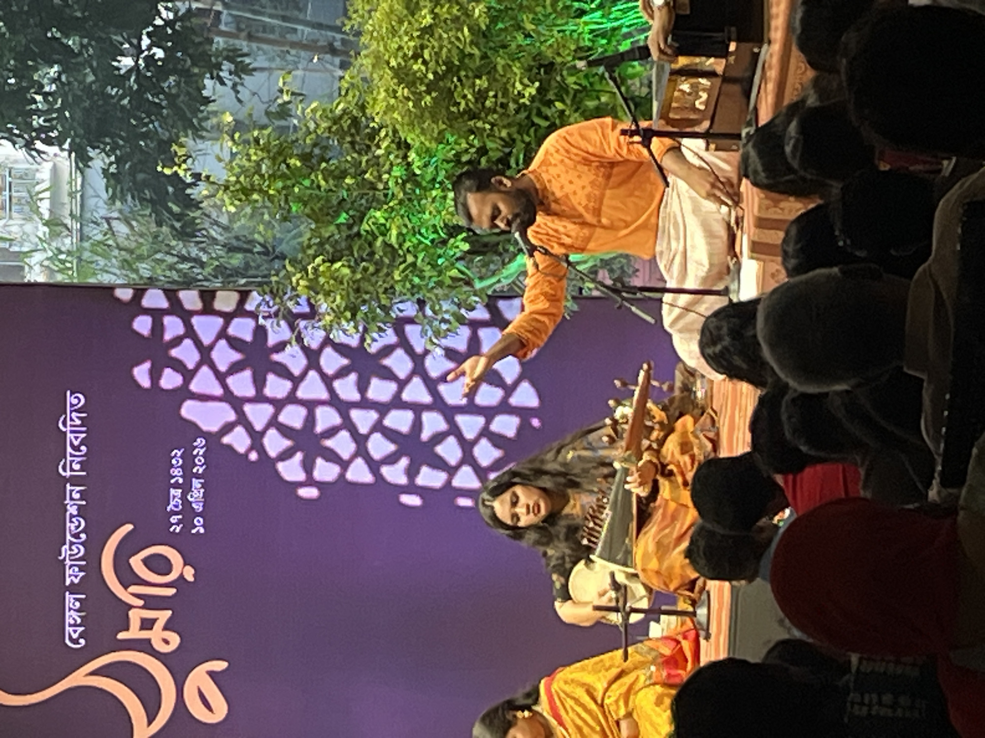 The image depicts a live musical performance. A man in an orange shirt and white pants is singing into a microphone, gesturing with his left hand. Seated below him, a woman is playing a stringed instrument, while another woman in a yellow garment is partially visible. A backdrop features an intricate geometric pattern and Bengali script. The performance is attended by an audience, whose heads and shoulders are visible in the foreground. The location appears to be outdoors, with trees and possibly a building visible in the background. The city is unknown.