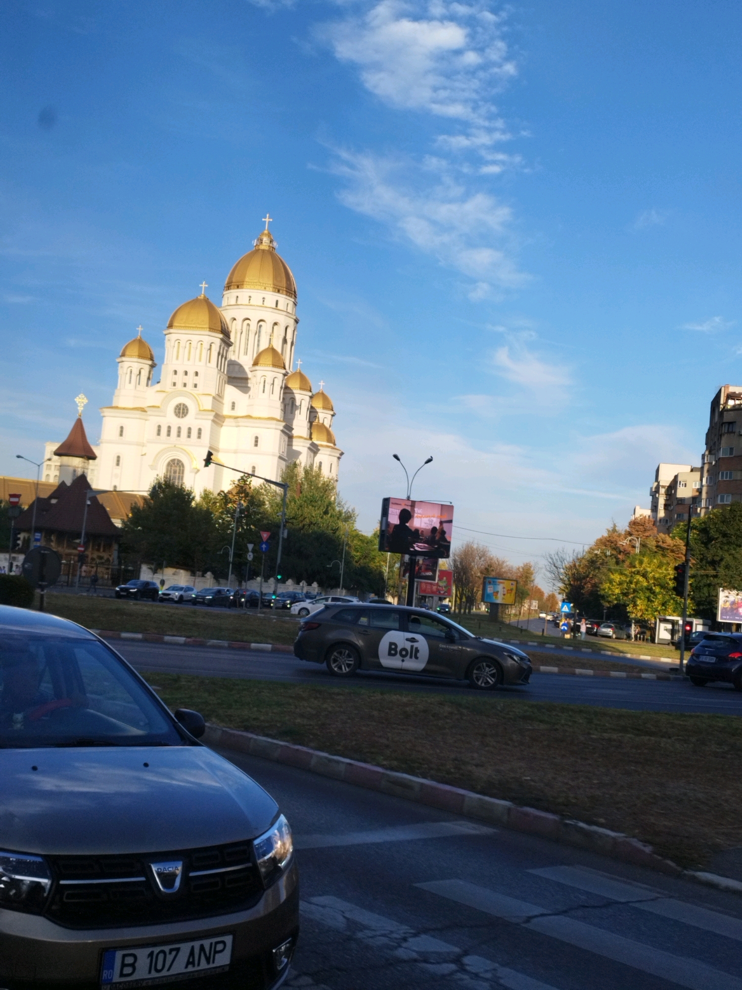 The image captures a street scene in Bucharest, Romania, with the imposing Metropolitan Cathedral of Bucharest as the dominant landmark. The cathedral, a grand structure with multiple golden domes and white facades, stands tall against a clear blue sky with wispy clouds. In the foreground, a Dacia car with the license plate "B 107 ANP" is partially visible. A Bolt ride-sharing vehicle is also seen driving on the street. Traffic lights and street lamps are present, indicating a city environment. Surrounding the cathedral are trees with autumn foliage and apartment buildings lining the street. Billboards are also visible in the background. The scene suggests a typical day in the city with ongoing traffic and urban activity.