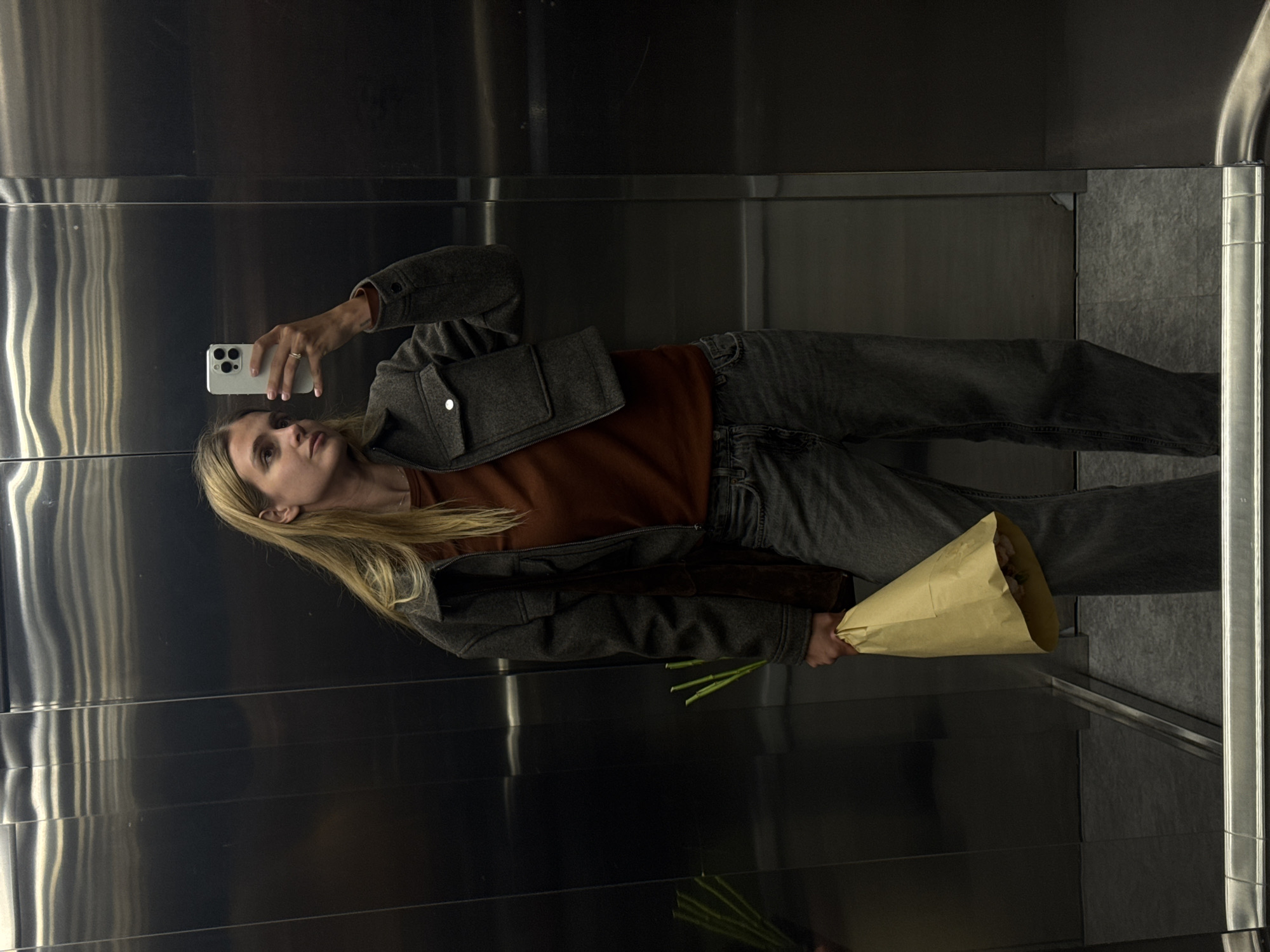 A woman stands in an elevator, holding a bouquet of flowers in a paper cone and taking a selfie with her white smartphone. She is wearing a brown top, a grey jacket, and grey jeans. The elevator has mirrored or metallic walls. The location appears to be Tbilisi, Georgia.