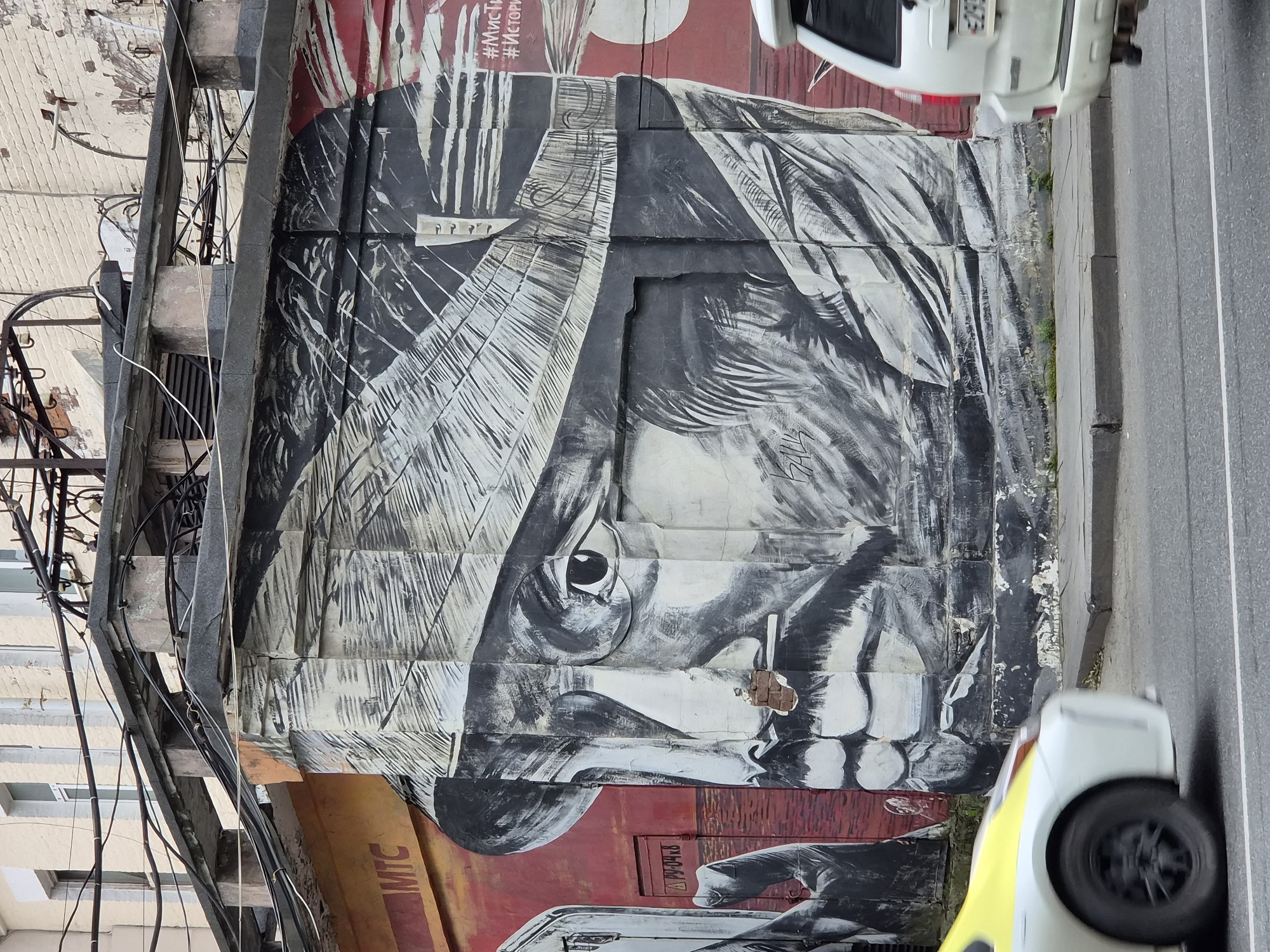 The image depicts a large mural on the side of a building in Vladivostok, Russia. The mural features a black and white portrait of a man wearing a hat, rendered in a dramatic, sketch-like style. Parts of the mural are peeling, revealing the underlying wall and brickwork. To the left of the mural, there is an orange section with the logo "MTC." To the right, there is a red section with white text that appears to be hashtags. A white vehicle with a yellow hood is blurred in the foreground on the left, suggesting movement. A white SUV is visible on the right, also slightly blurred. The mural is situated above a sidewalk and a street with cars. Overhead, electrical wires and conduits crisscross the scene, attached to the building and poles. The architecture suggests an urban environment, with visible windows and brickwork on the upper floors of the building.
