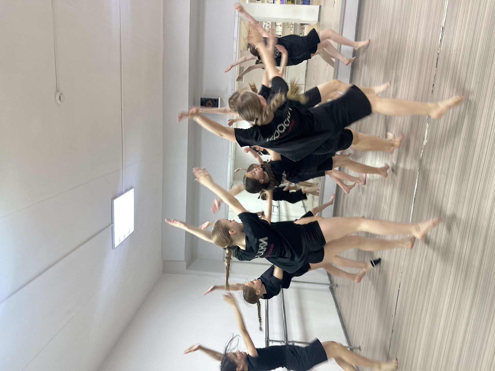 A group of at least 9 young dancers, all wearing matching black T-shirts and shorts, are captured mid-motion in a dance studio. The studio is located in Votkinsk, Russia. The dancers are positioned at varying points in a leaping movement. Their arms are outstretched, and their legs are extended in a coordinated fashion. The studio features a light-colored, possibly grey, floor with subtle linear patterns, a white ceiling, and a wall-sized mirror reflecting the dancers and the room's interior. A ballet barre is visible against the back wall of the studio. The space appears well-lit, with natural light coming from a rectangular window high on the wall.
