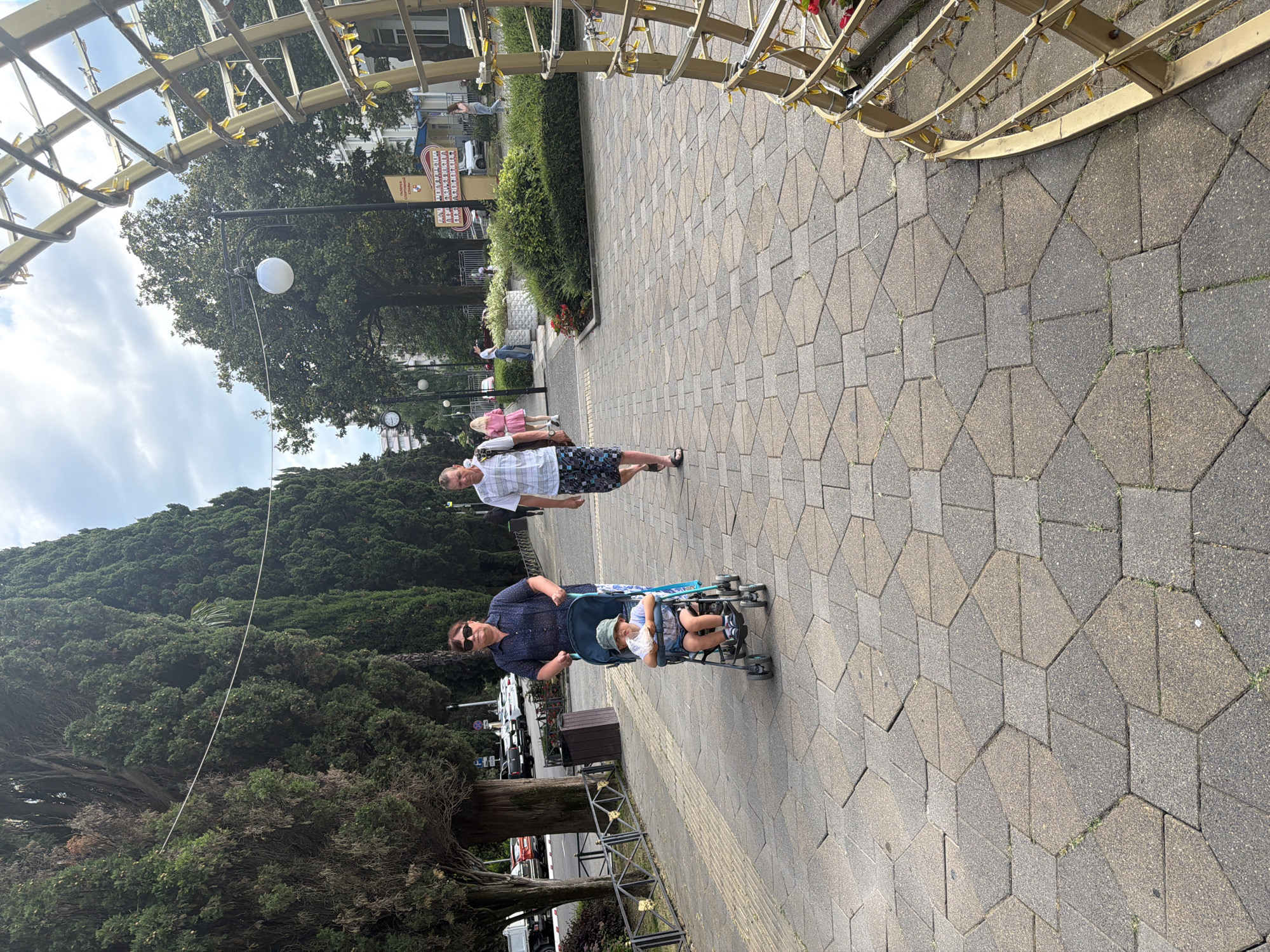 In Sochi, Russia, a paved walkway made of hexagonal and square stones features three people and a stroller. A woman, wearing sunglasses and a dark patterned shirt, pushes a stroller with a young child inside. The child, wearing a light-colored hat and shorts, is seated in the stroller, holding something white. A man in a patterned short-sleeved shirt and patterned shorts walks away from the camera, passing the woman and stroller. The walkway is bordered by tall trees on the left and a decorative metal structure with lights on the right. In the background, there are more trees, buildings, and a parked car. The sky is overcast.